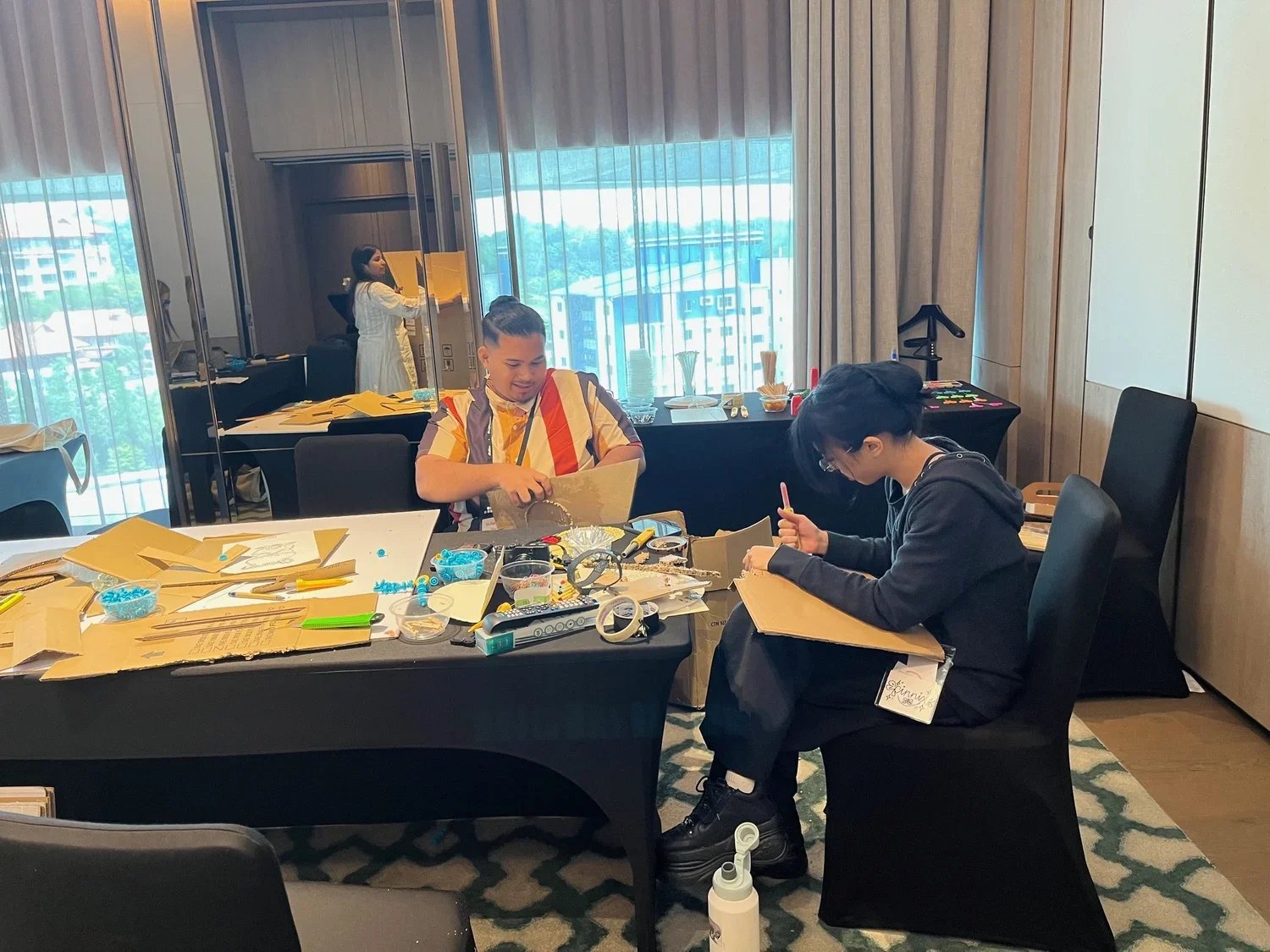 Two workshop participants sit at a table covered with cardboard, markers, tape, and craft supplies, working on hands-on design projects while another participant works in the background.