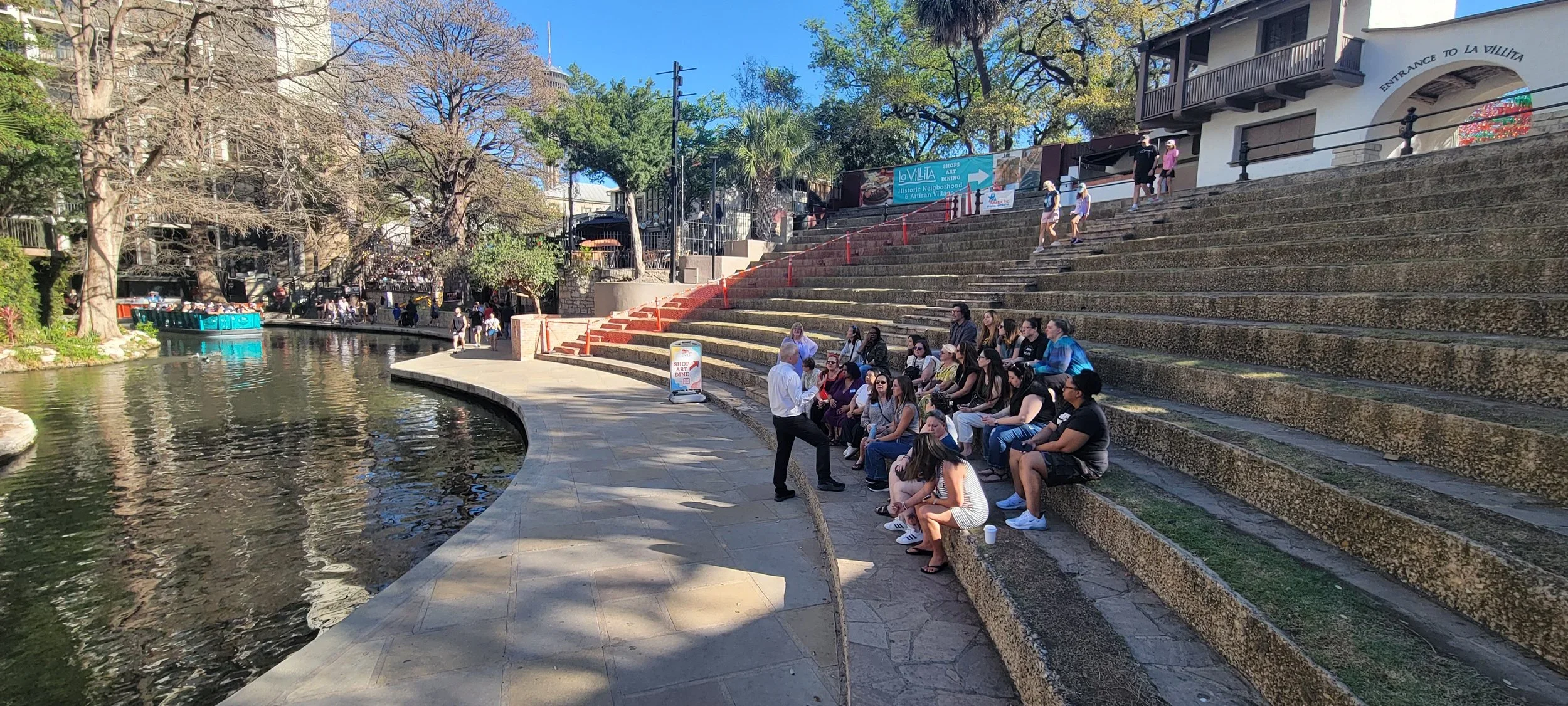 A group of people sits on tiered stone steps beside a canal during a guided tour. Trees, historic buildings, and a riverwalk create a scenic urban gathering space.