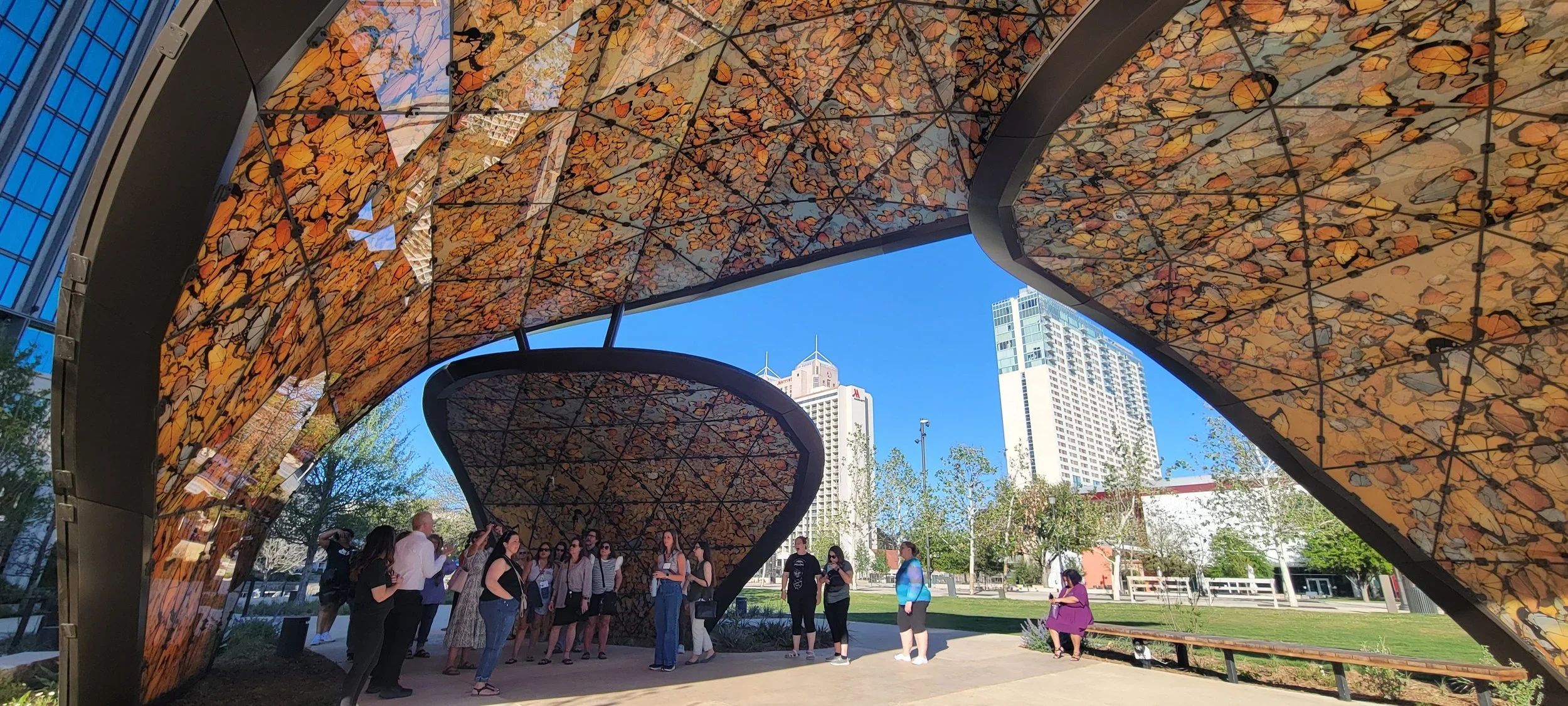 Visitors gather beneath a sculptural canopy with a colorful leaf-patterned underside in an urban park. Modern high-rise buildings and landscaped green space surround the pavilion.