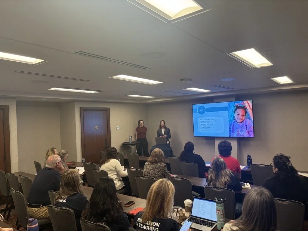 Attendees sit in a conference room during a presentation, watching two speakers at the front of the room. A large screen displays presentation slides as participants listen from their seats.