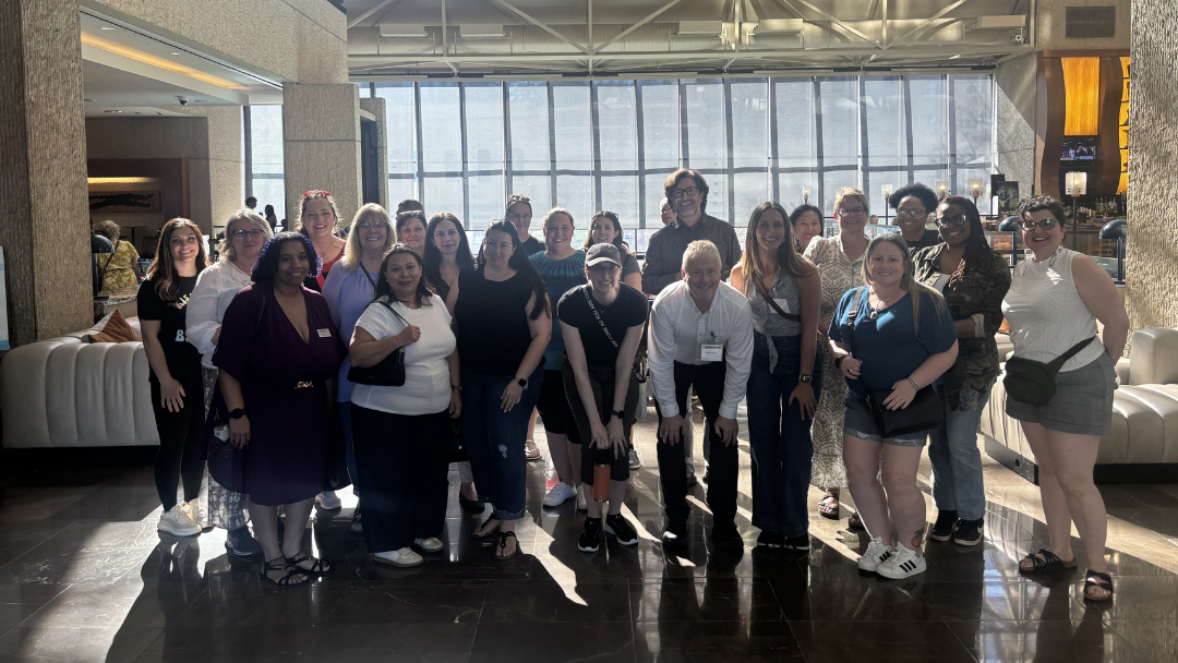 A large group poses for a photo in a hotel lobby with floor-to-ceiling windows. Sunlight fills the space as attendees smile together during a conference or group gathering.