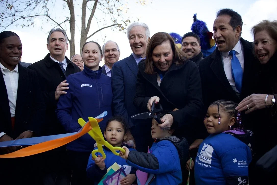 Children and community leaders celebrate a ribbon-cutting ceremony outdoors, using oversized scissors to open a new early childhood education facility while supporters gather around.