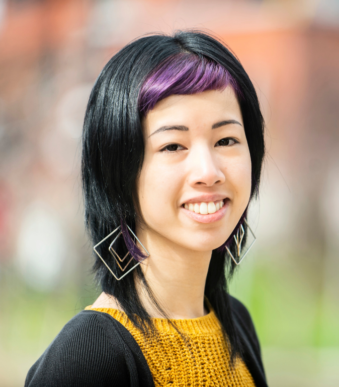 Professional headshot of a smiling woman with black hair and purple bangs, wearing geometric earrings and a mustard-yellow sweater, photographed outdoors with a softly blurred background.