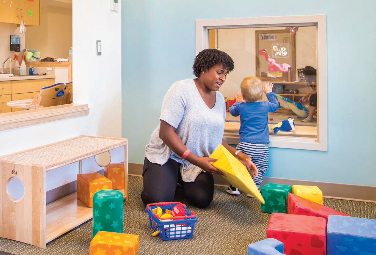A woman kneeling on a carpeted floor in a daycare or preschool setting, placing foam blocks around children who are playing. There are colorful foam blocks scattered on the floor, with some inside a blue toy basket. Behind a glass window, a child with blond hair and striped pants is reaching to touch the window, and another child and an adult are in an adjacent room playing.