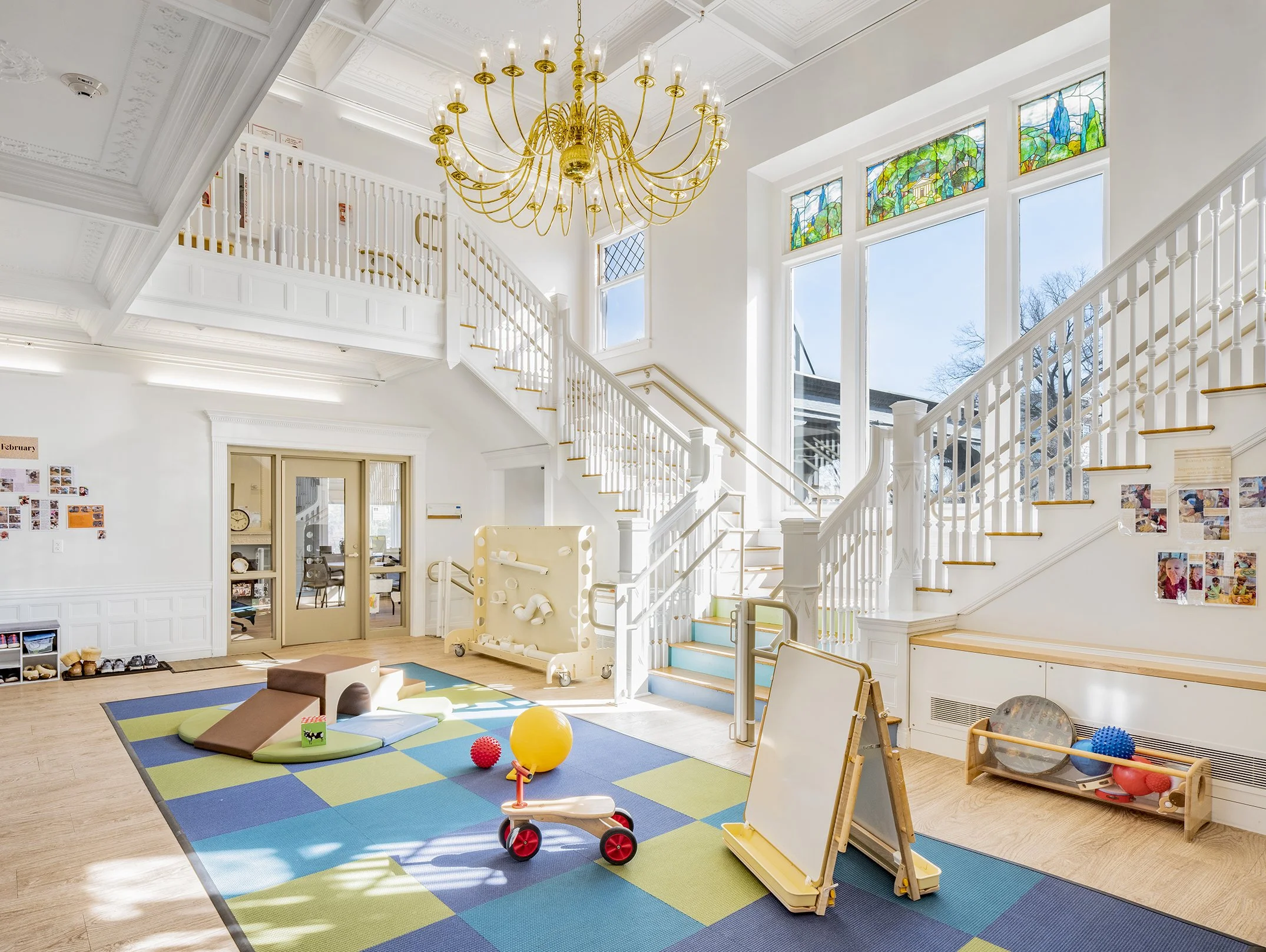 Bright, double-height early childhood learning space within a restored historic building, featuring a grand staircase, stained-glass windows, natural light, soft play equipment, and activity areas for young children.