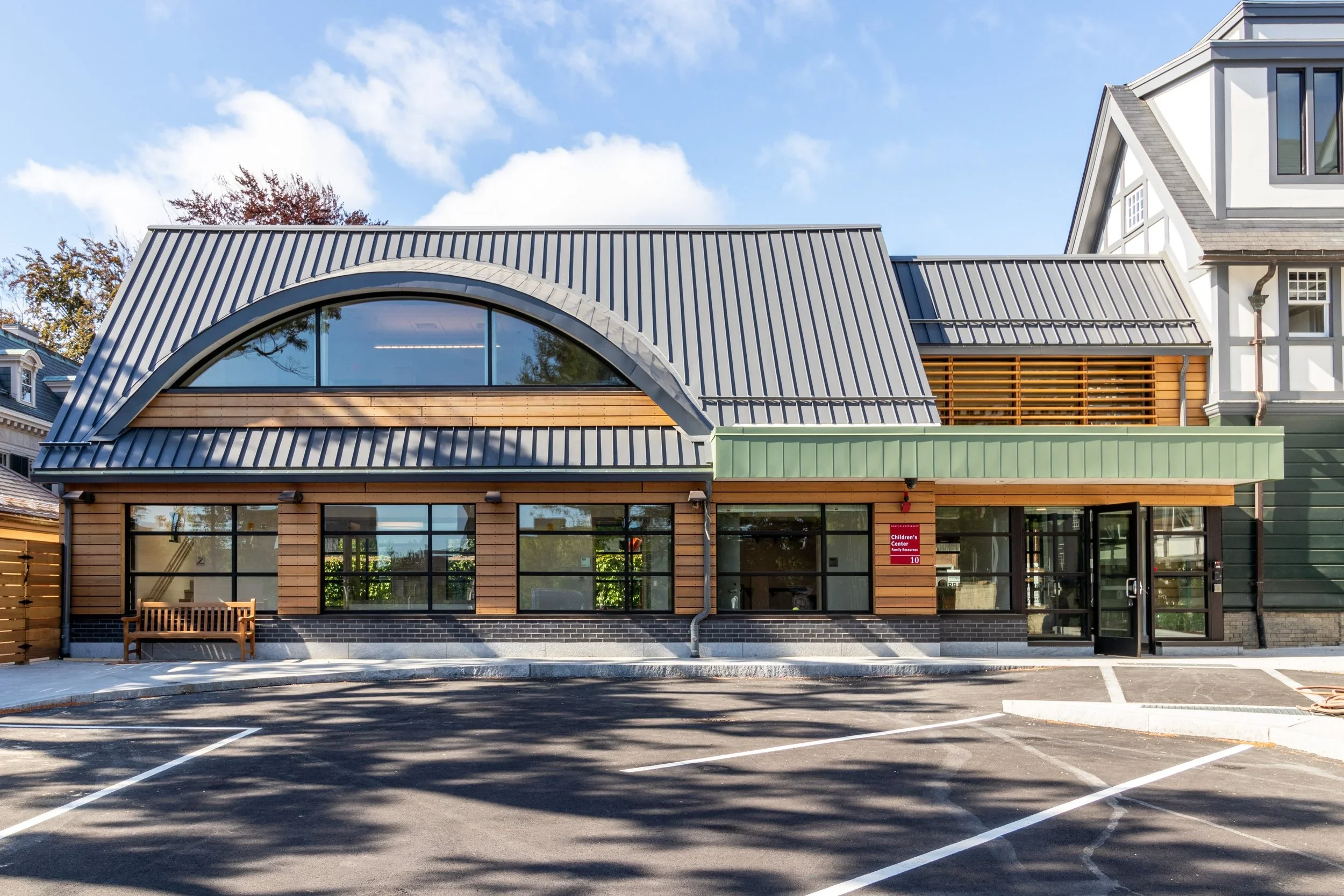 Exterior view of a children's center addition featuring wood and metal cladding, expansive windows, a curved roofline, and a contemporary design that complements the adjacent historic building.
