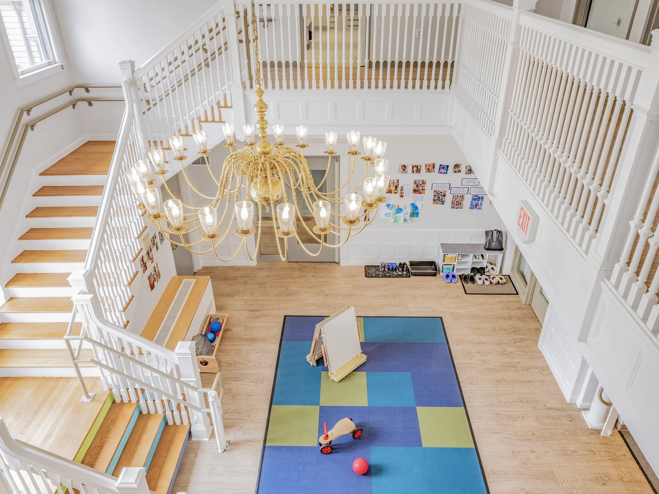 Bright, double-height early childhood learning space within a restored historic building, featuring a grand staircase, stained-glass windows, natural light, soft play equipment, and activity areas for young children.