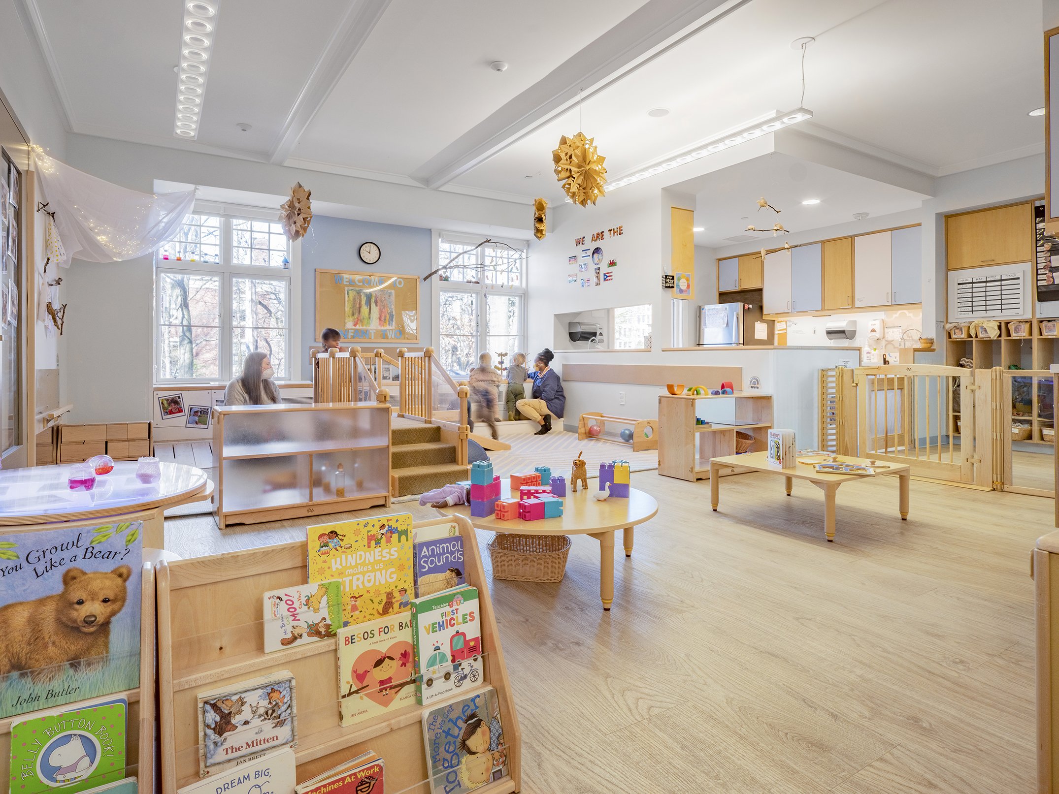 Infant classroom with child-scaled furnishings, books, sensory materials, and age-appropriate play areas. Large windows provide natural light, creating a warm environment for exploration and early learning.