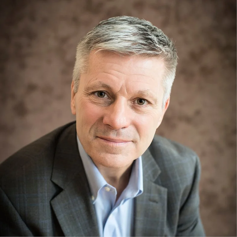 A person with short gray hair wears a dark blazer over a light blue collared shirt, looking toward the camera against a softly blurred brown background.