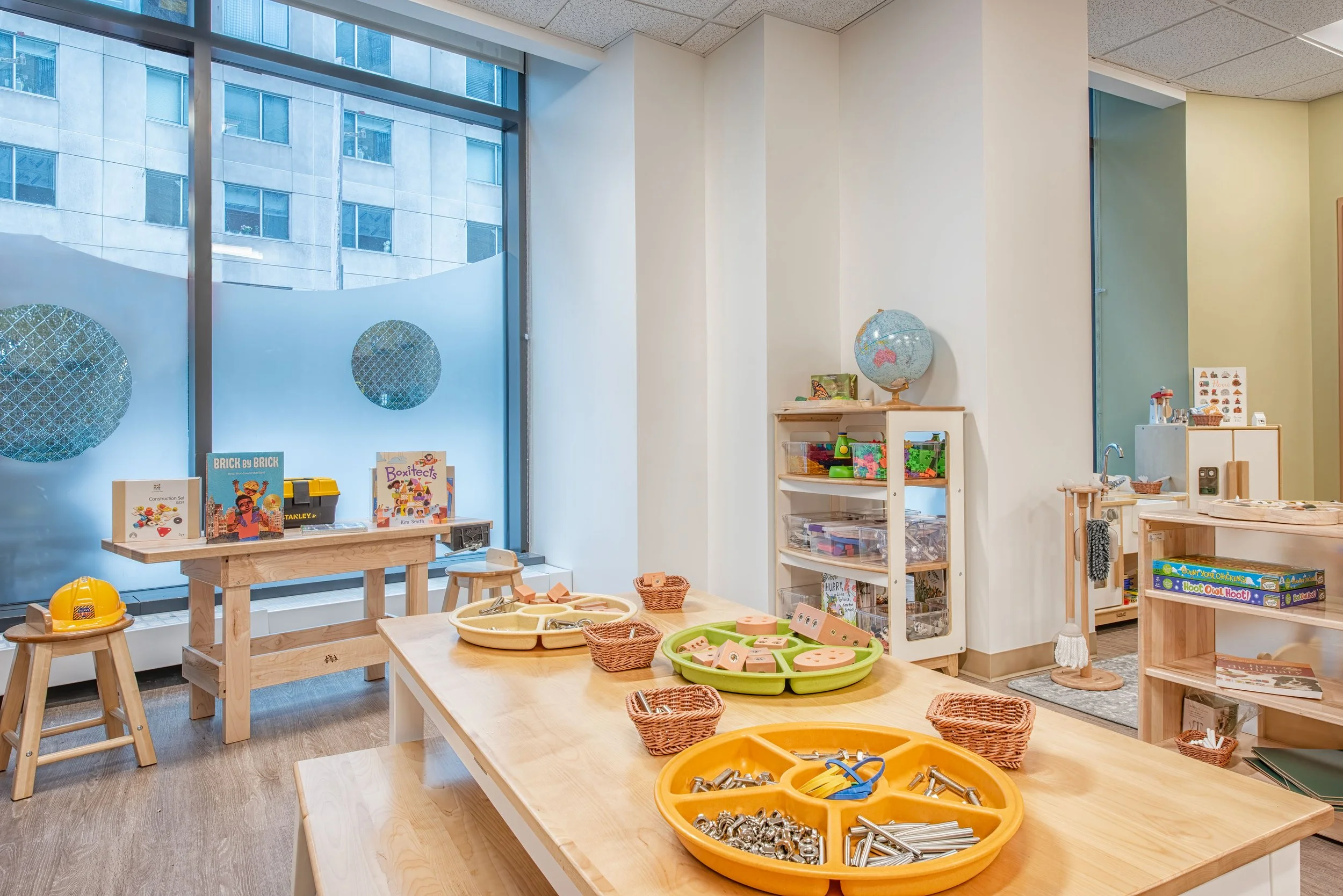 Early learning classroom with wood tables, construction-themed learning materials, open shelving, and large windows bringing natural light into the space.