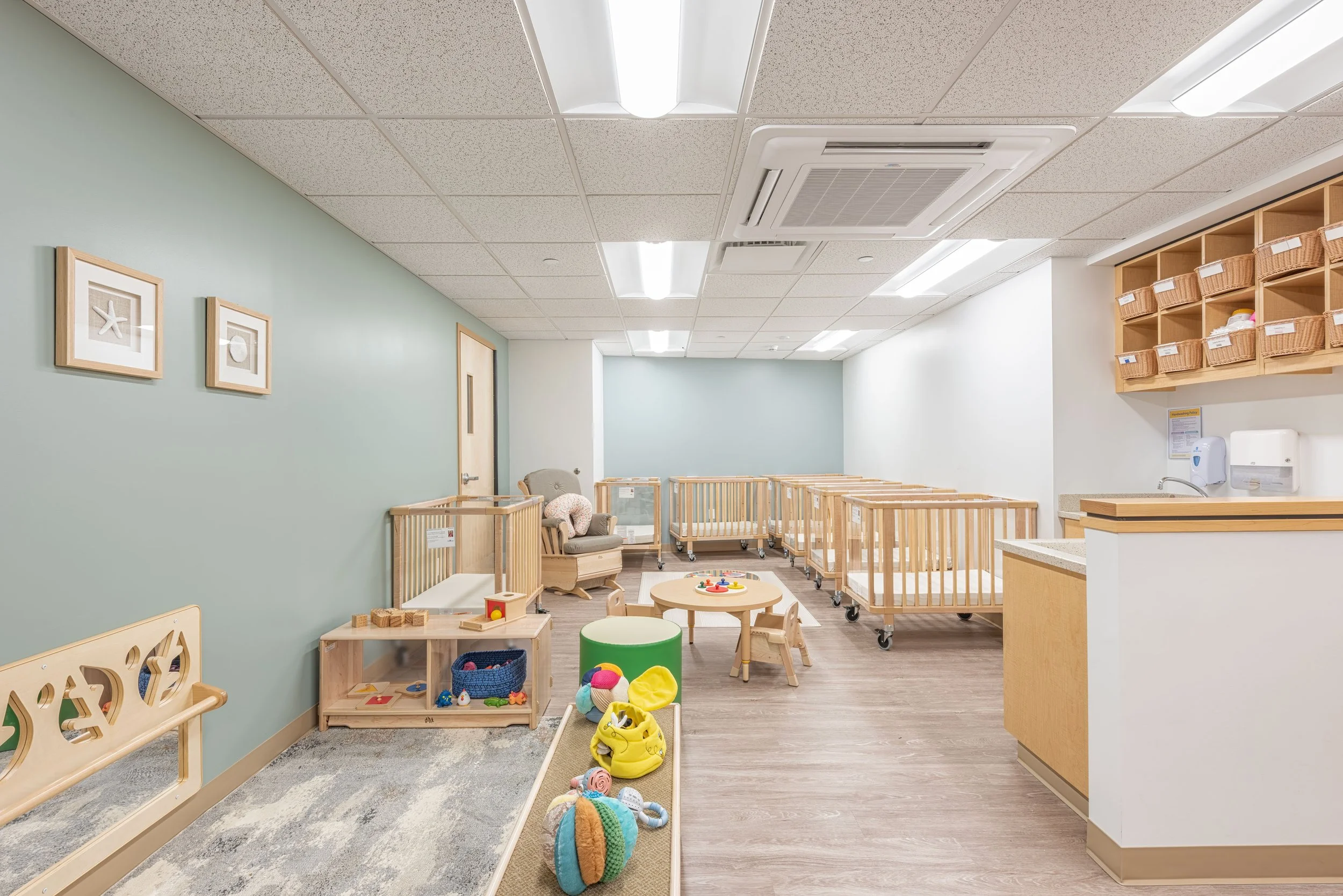 Infant classroom with cribs, child-sized furnishings, soft play areas, and muted green walls.