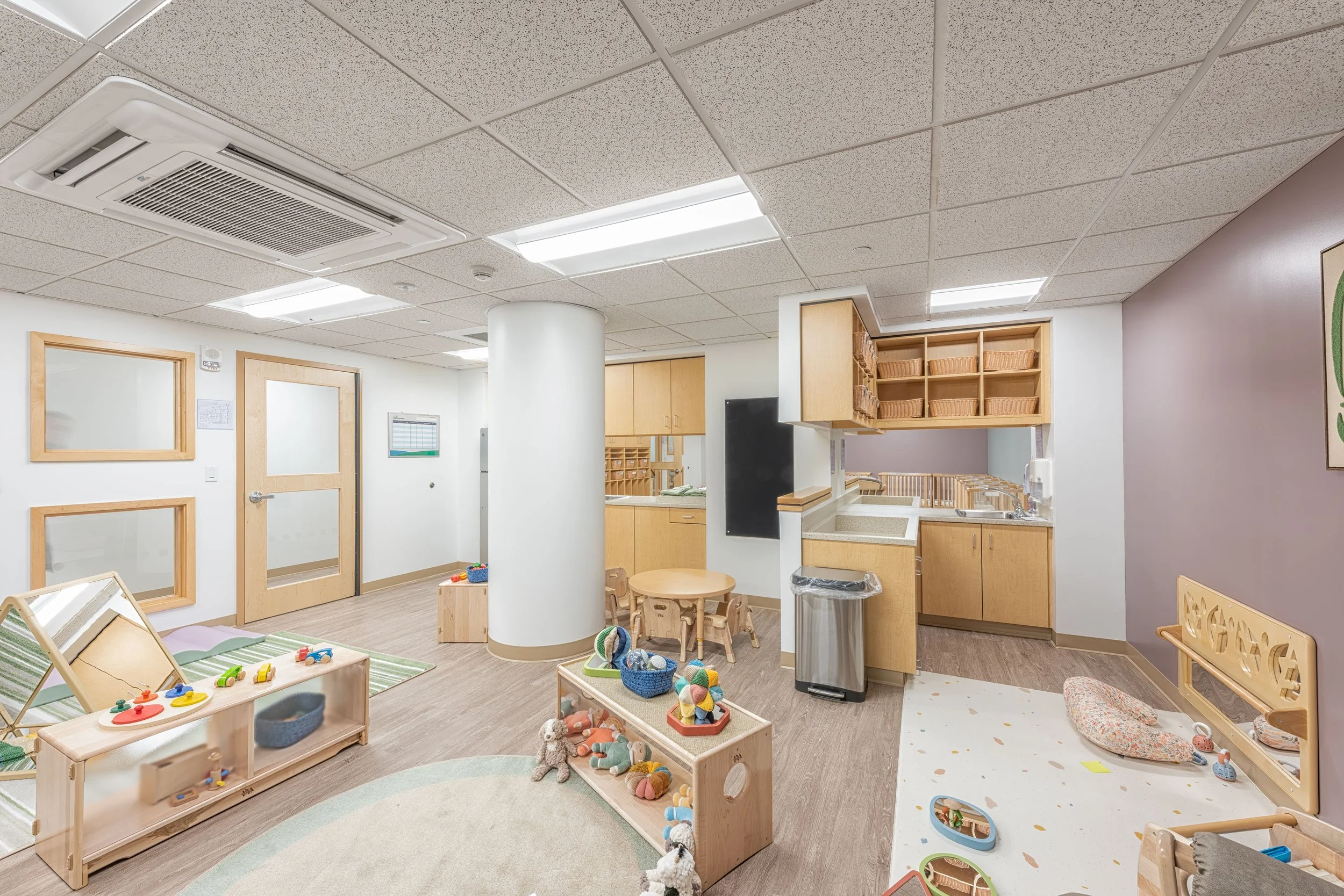 Infant classroom with low wood shelving, soft play areas, changing station, and interior windows supporting visibility between spaces.