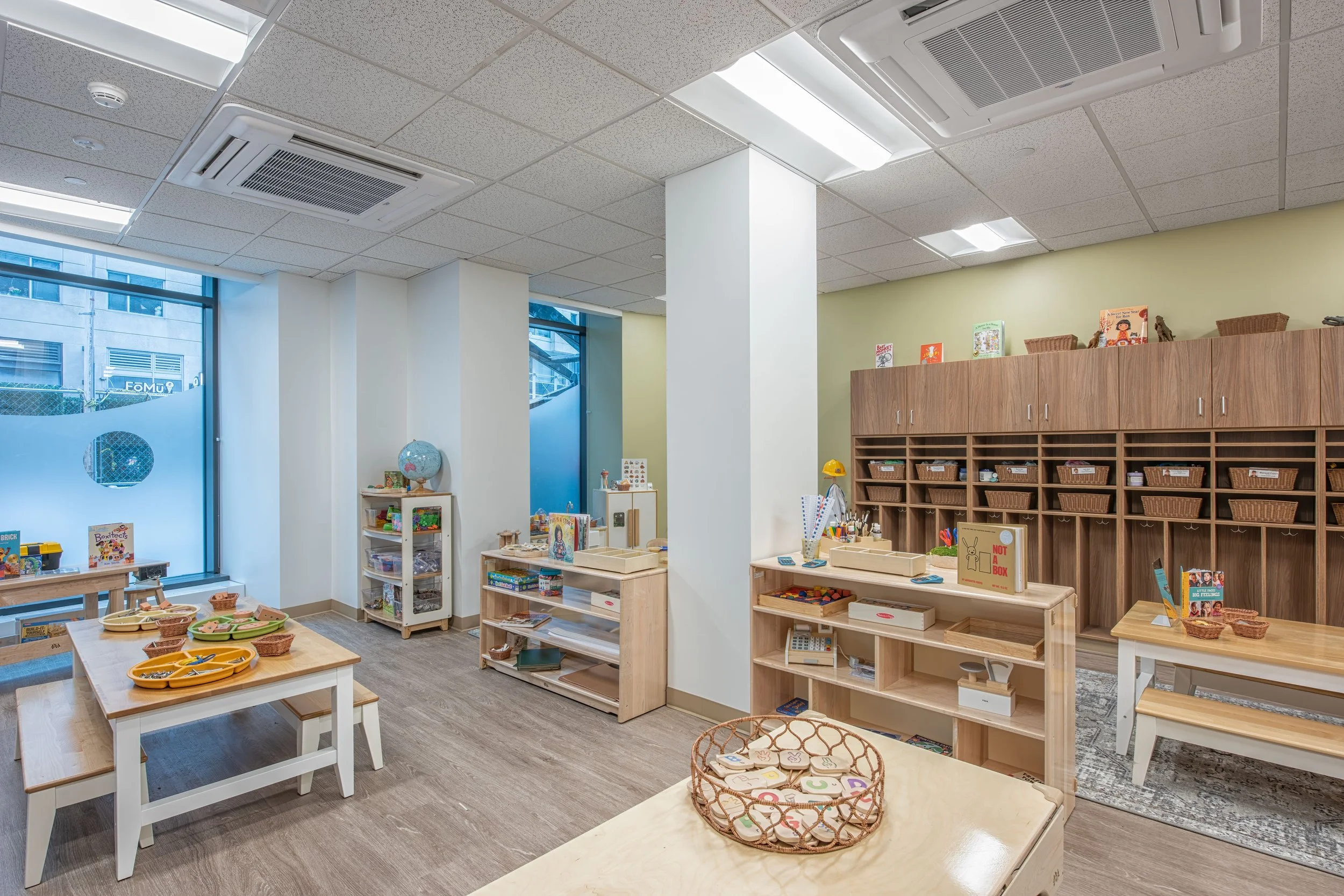Preschool classroom with child-sized tables, open wood shelving, activity materials, and large windows with patterned privacy glazing.