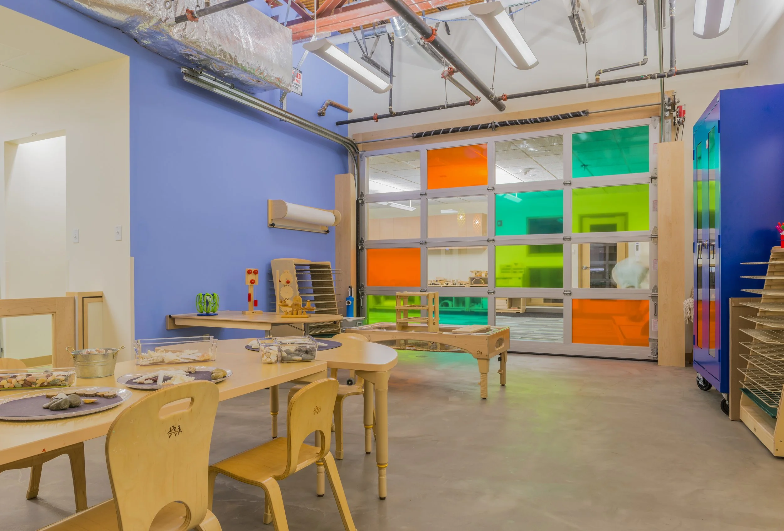 Early learning STEAM classroom with child-sized tables, hands-on materials, blue walls, and a garage-style door with colorful translucent panels.