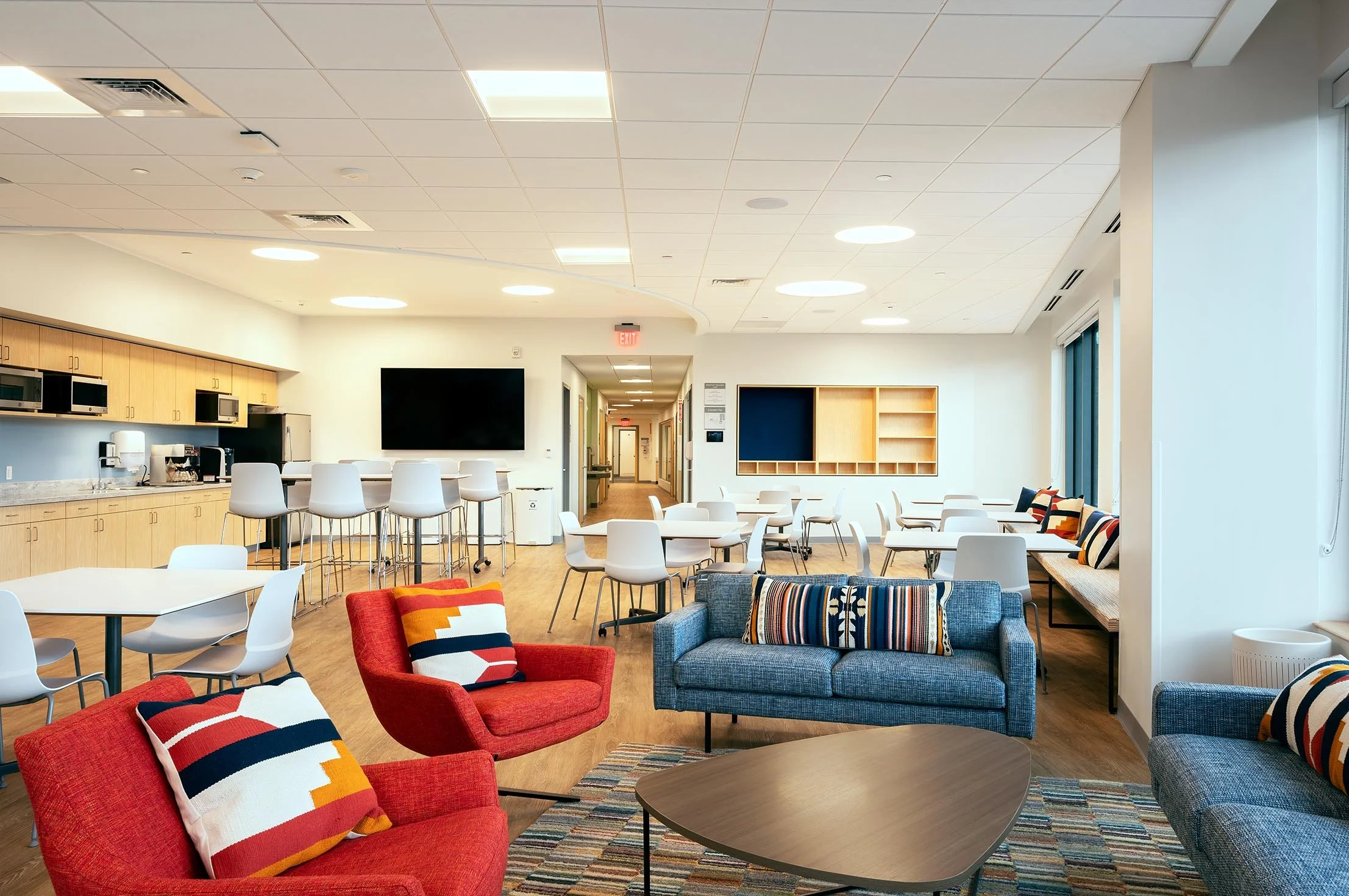 Staff lounge with kitchen, dining tables, colorful lounge seating, and large windows providing natural light.