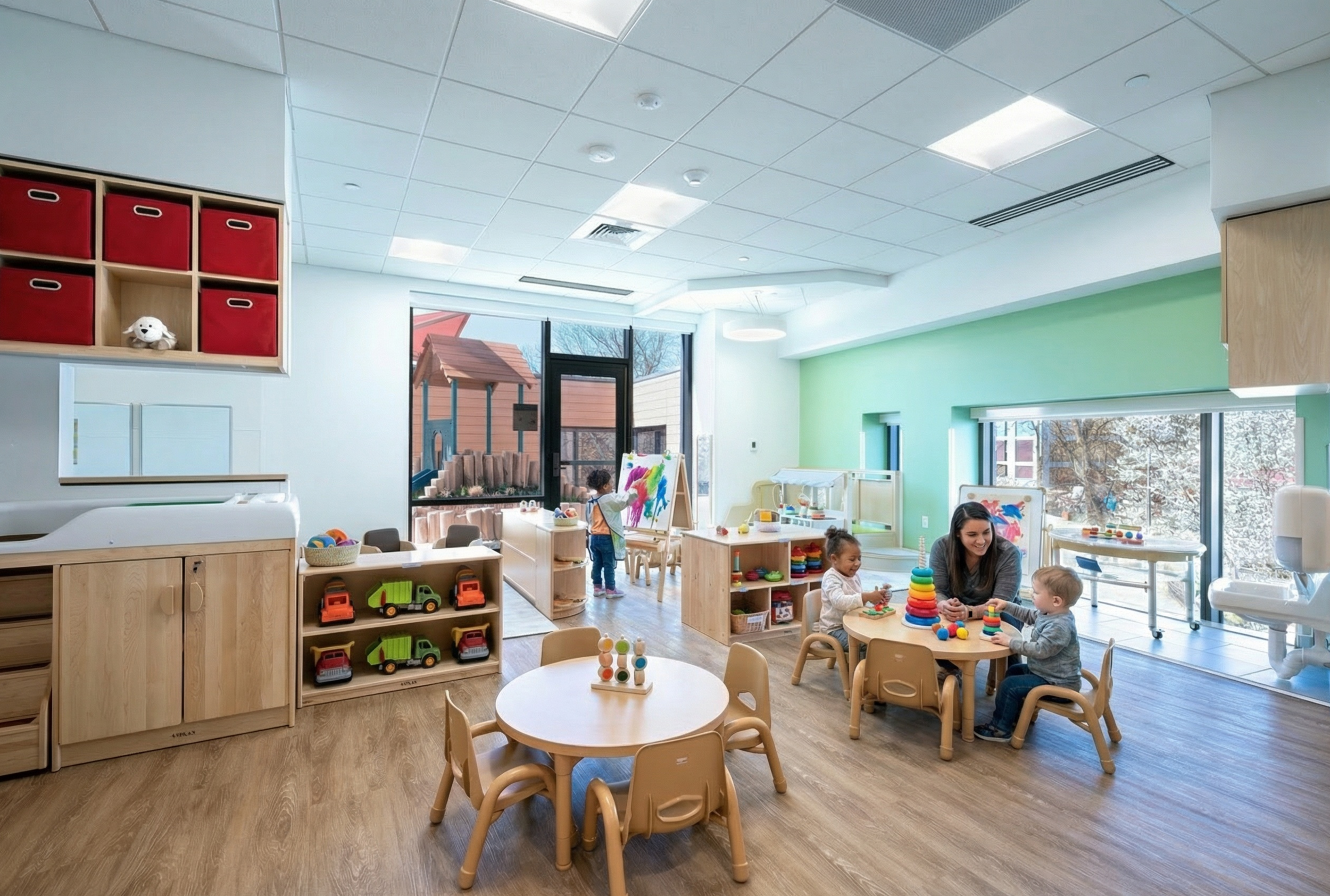 Children and a teacher participate in classroom activities in a light-filled toddler learning environment with child-sized furniture, open shelving, large windows, and direct access to an outdoor play area.
