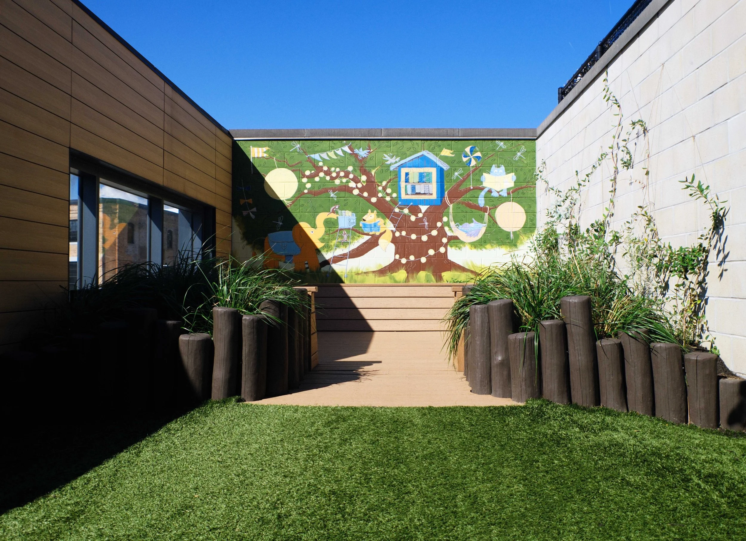 Outdoor play space with artificial turf, planted areas, natural wood elements, and a colorful mural.