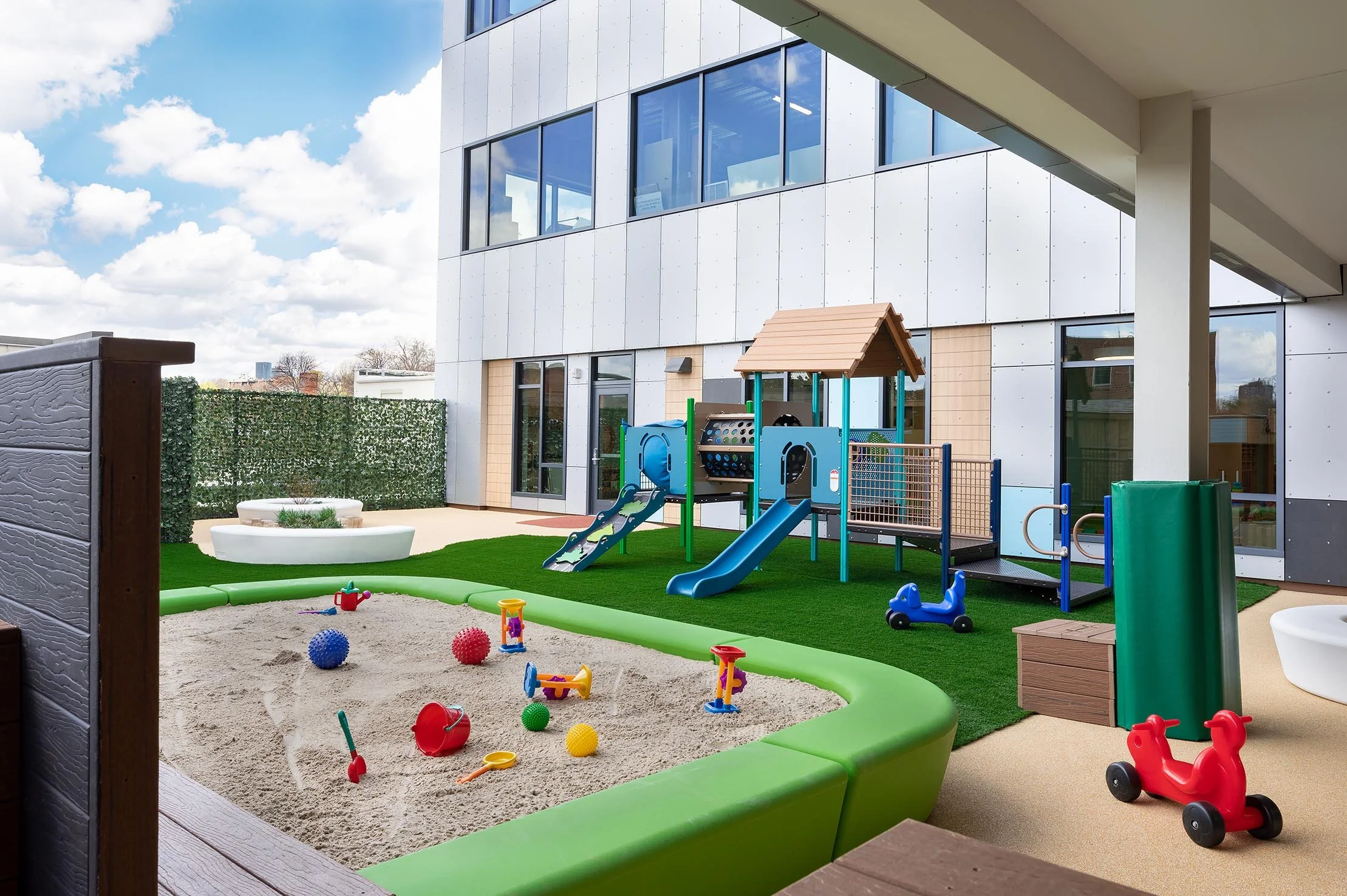 Early childhood playground with a large sandbox, play structure, slide, artificial turf, and riding toys.