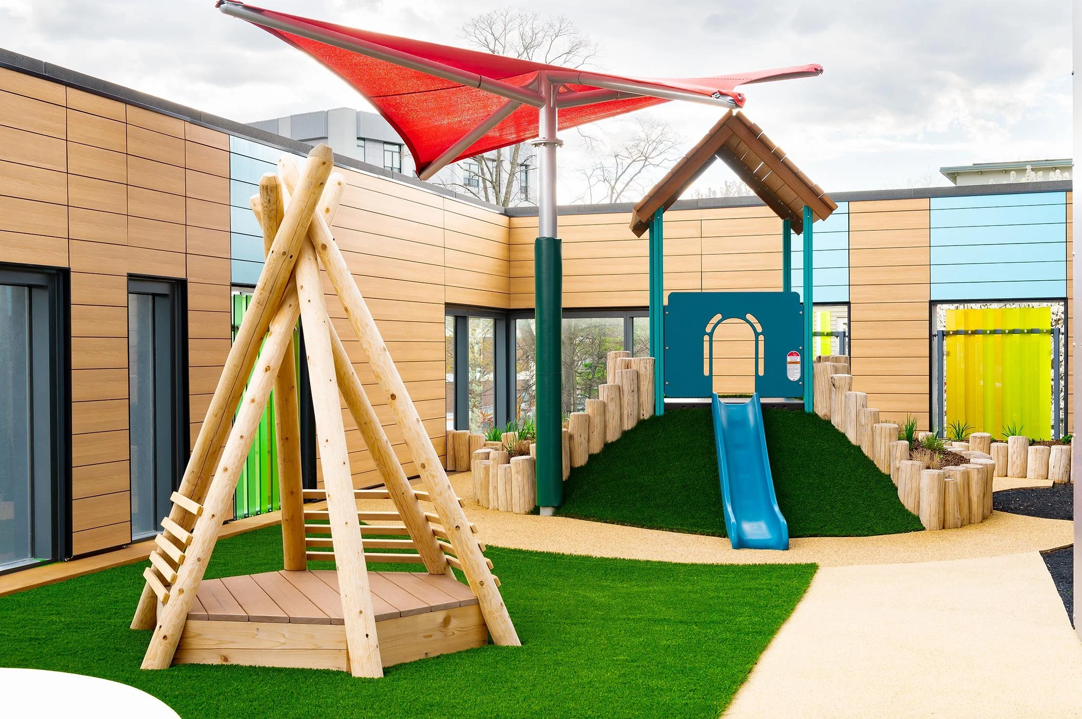Outdoor play area with a wood climbing structure, turf-covered hill and slide, shade canopy, and natural wood play elements.