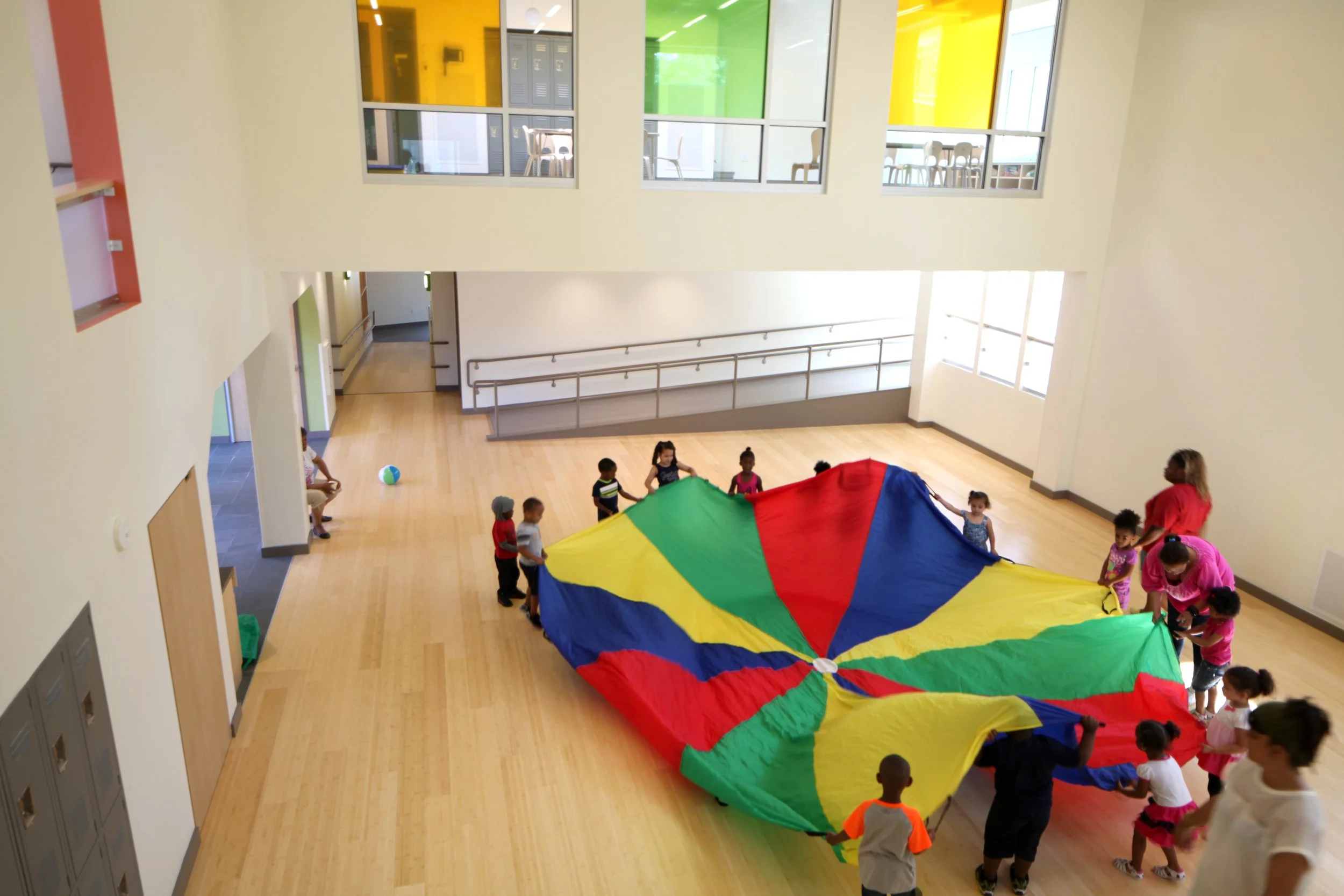 Children playing with a colorful parachute in a large, light-filled indoor multipurpose space at Nurtury Learning Lab.