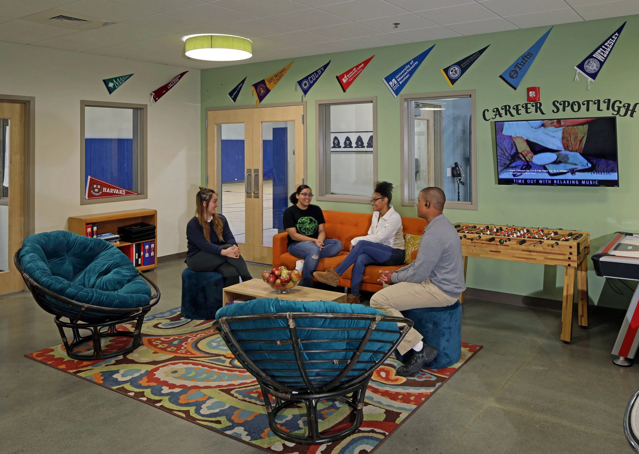 Students and adults gather in a colorful lounge with casual seating, a foosball table, and college pennants displayed along the wall.