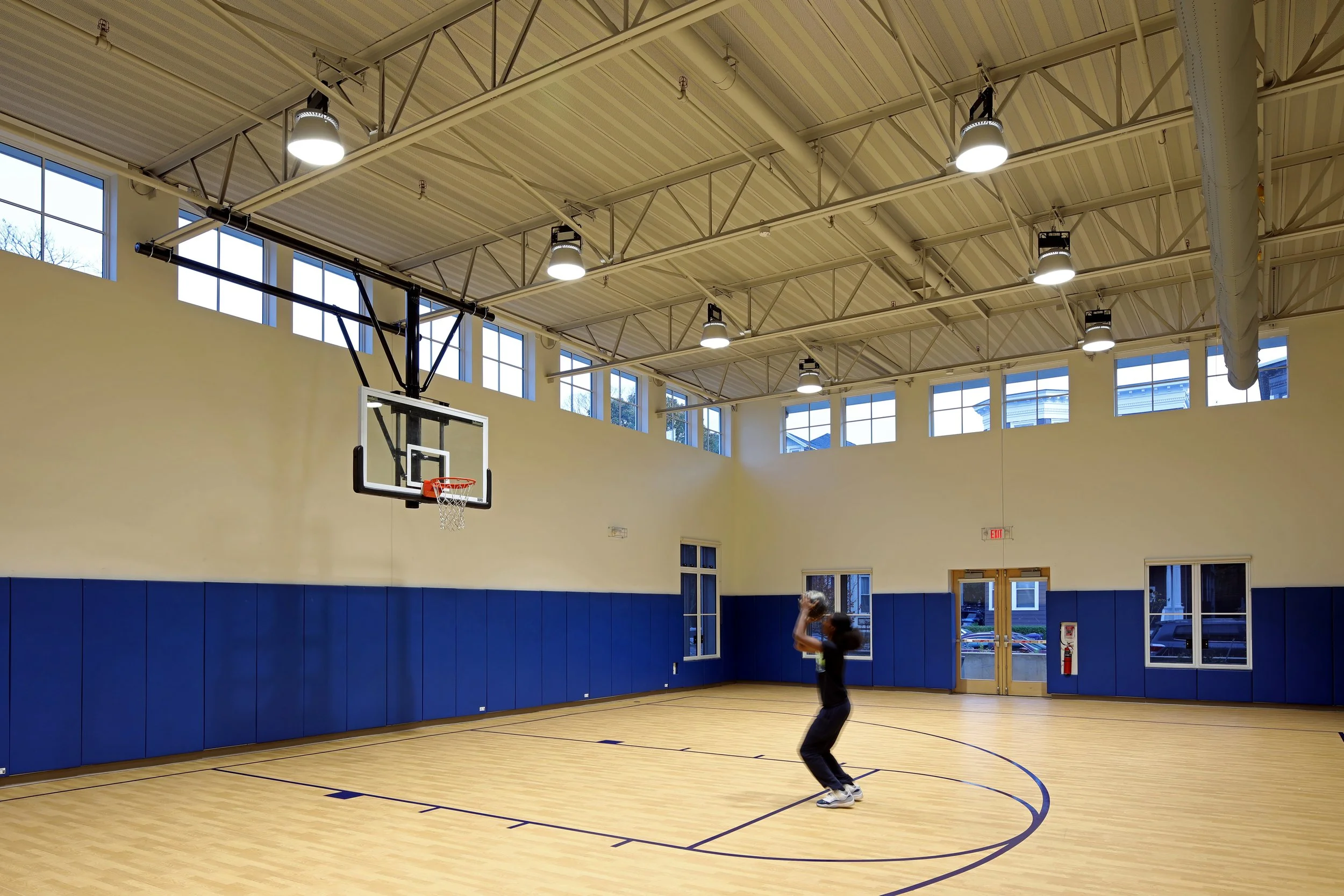 Student playing basketball in a bright gymnasium with a wood court, blue wall padding, and high ceilings.