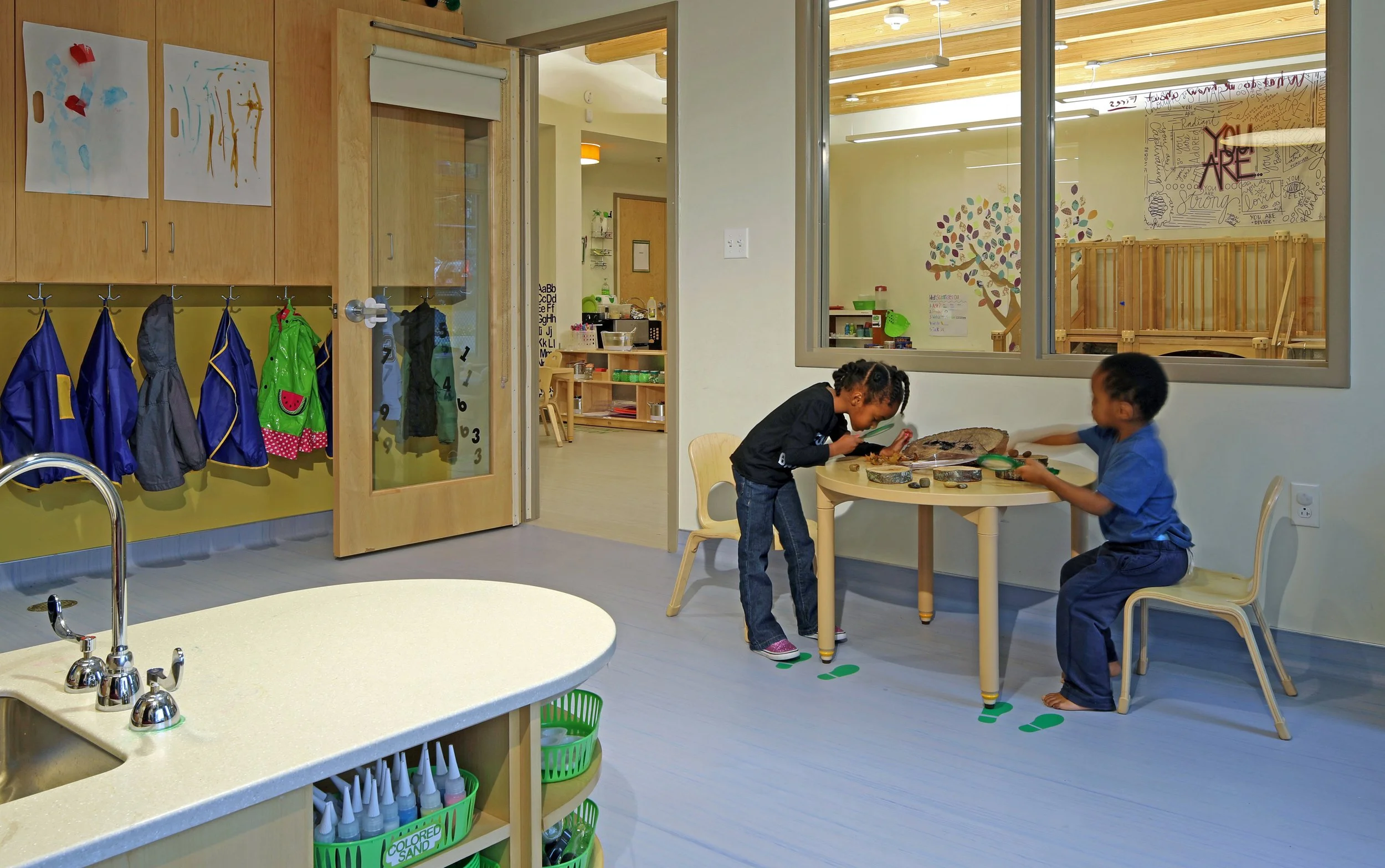 Two children explore natural materials at a classroom activity table beside interior windows connecting adjacent learning spaces.