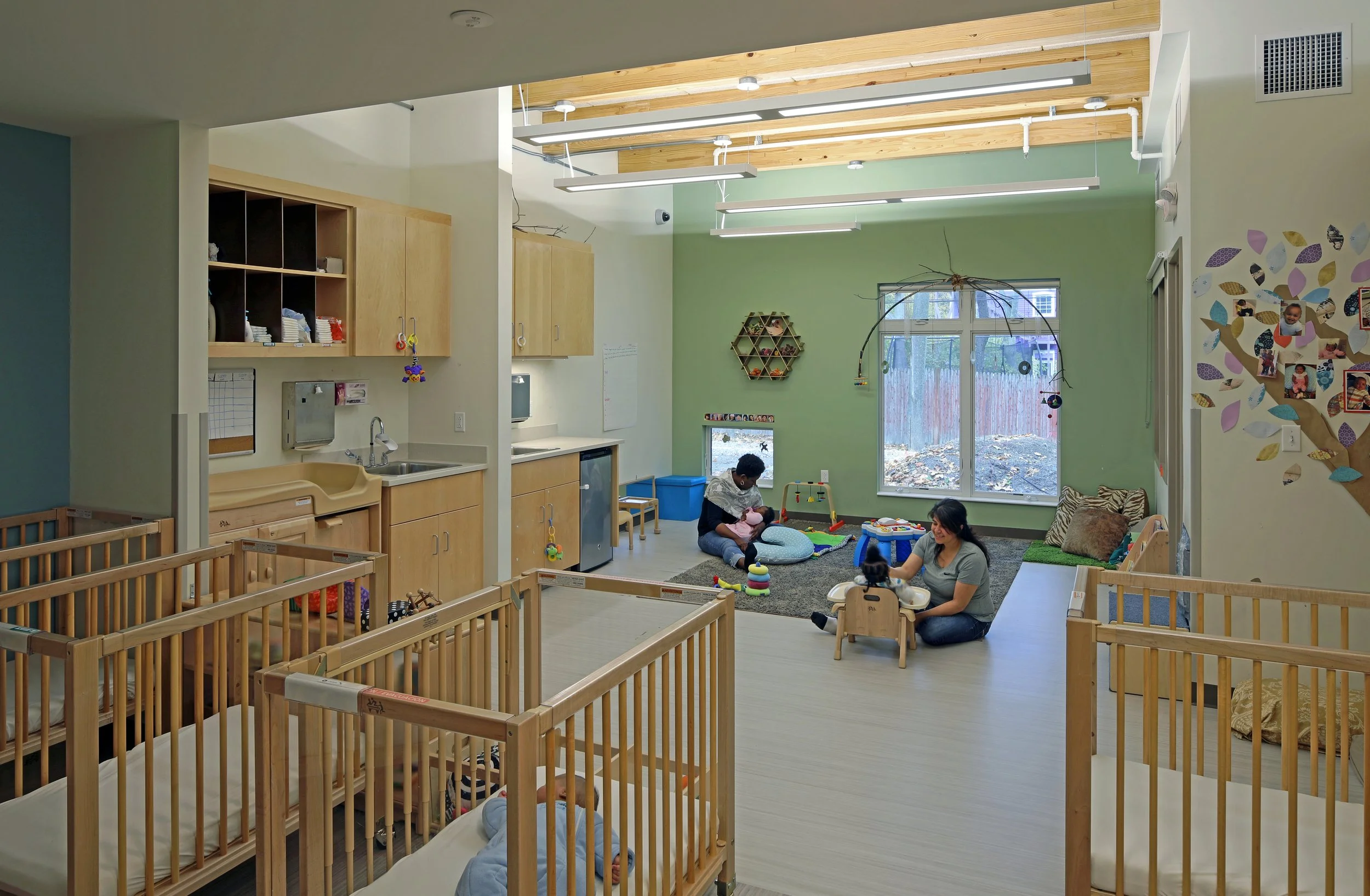 Bright early learning classroom with cribs, built-in cabinetry, play areas, and caregivers interacting with young children.