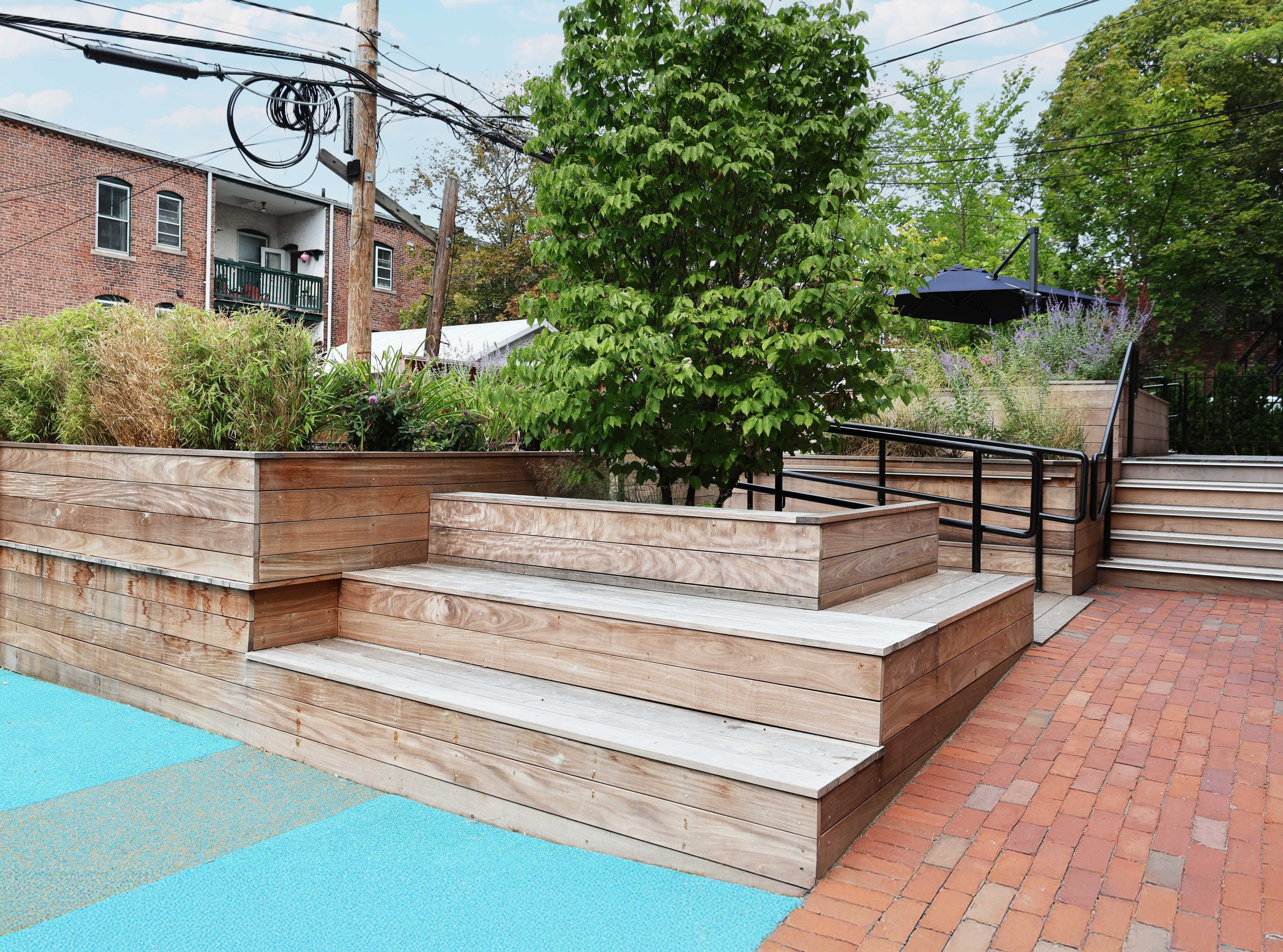 Therapeutic outdoor space with tiered wood seating, raised planters, mature trees, an accessible ramp, and brick paving.