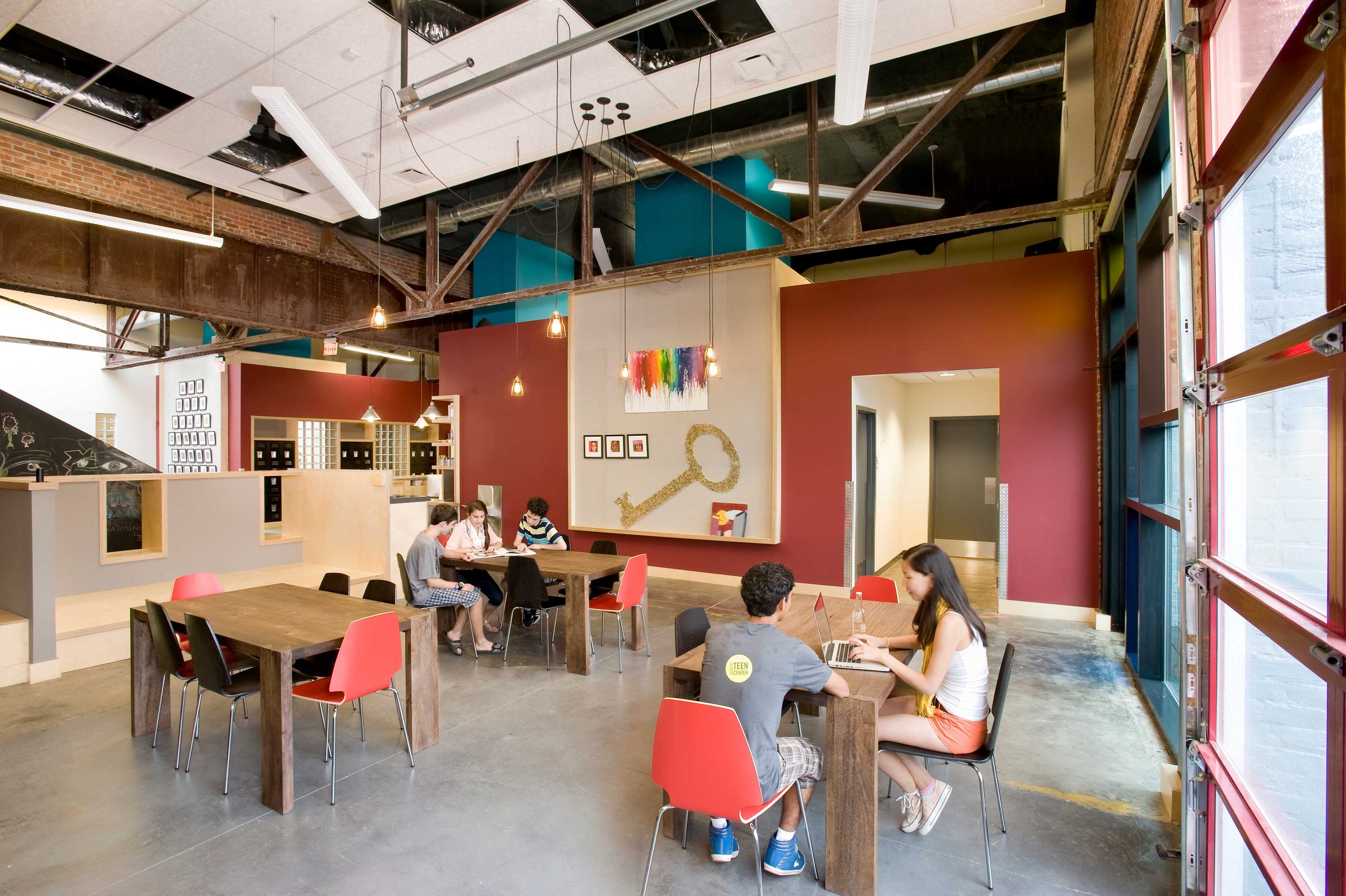 Teens study and socialize at tables in an open multipurpose space featuring exposed structural elements, colorful walls, and flexible furnishings.