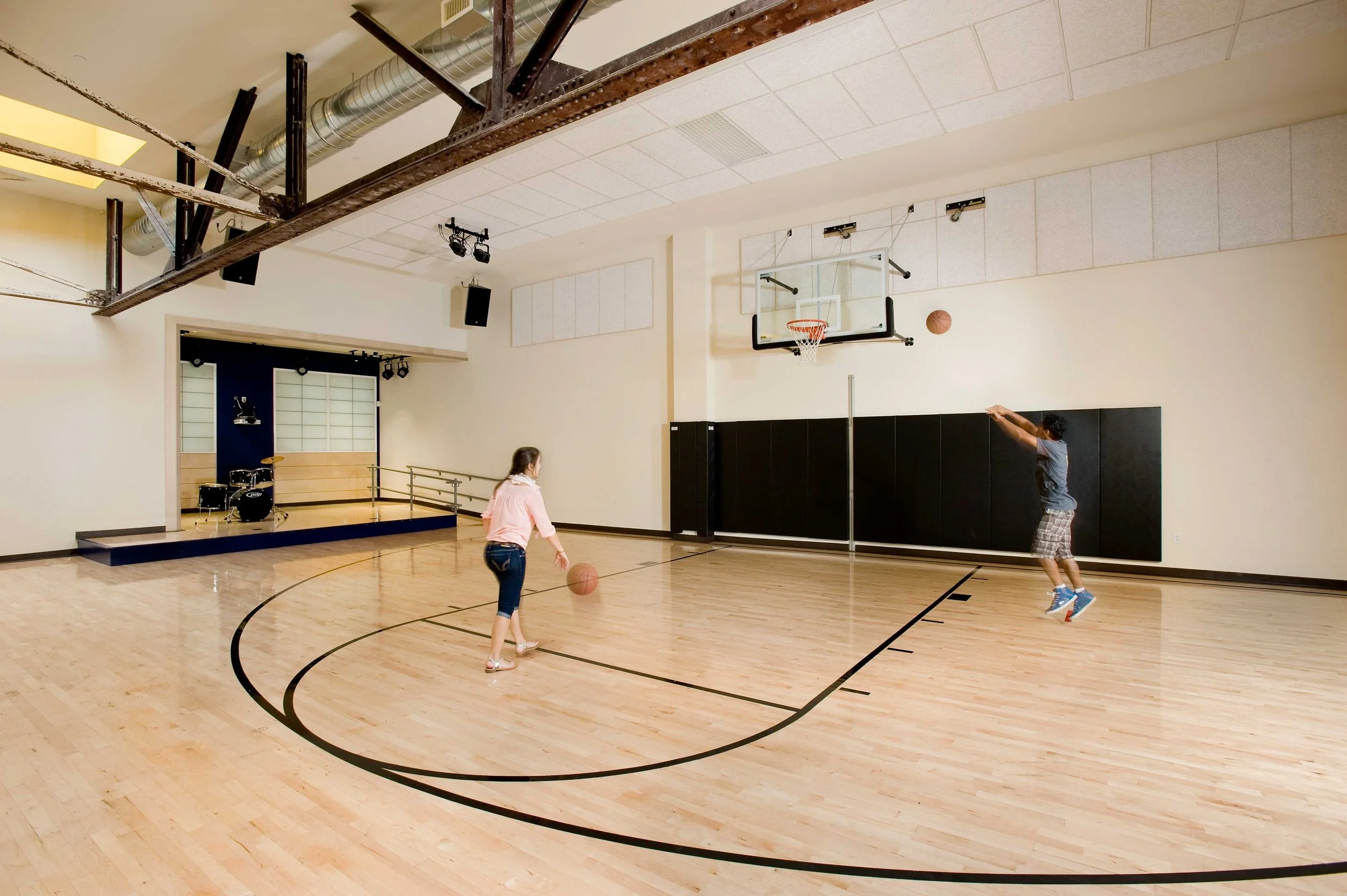 Teens play basketball in a multipurpose gym with a wood court, basketball hoop, wall padding, and a small performance stage.