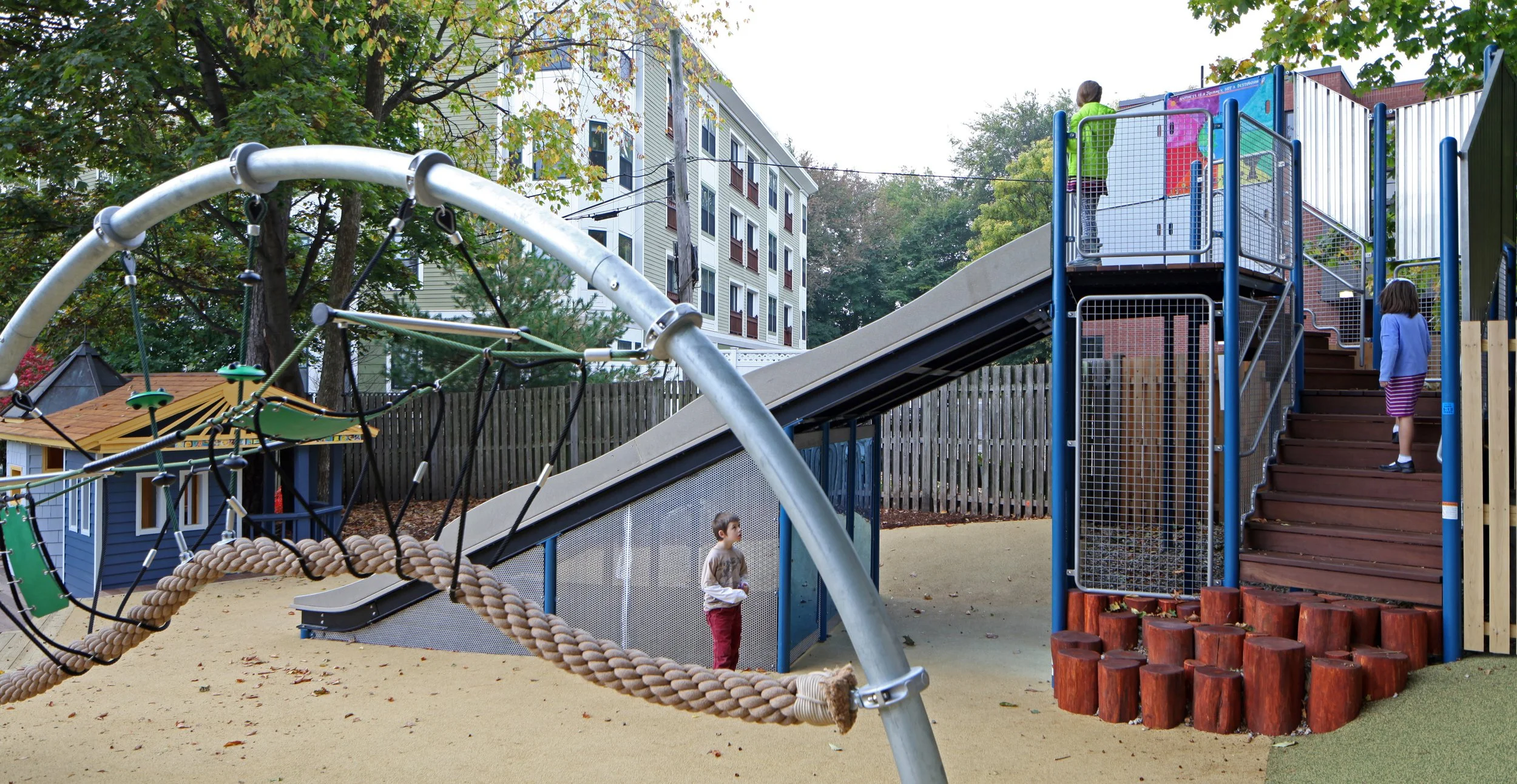 Children exploring a playground with a large slide, rope climbing equipment, and wooden steps.