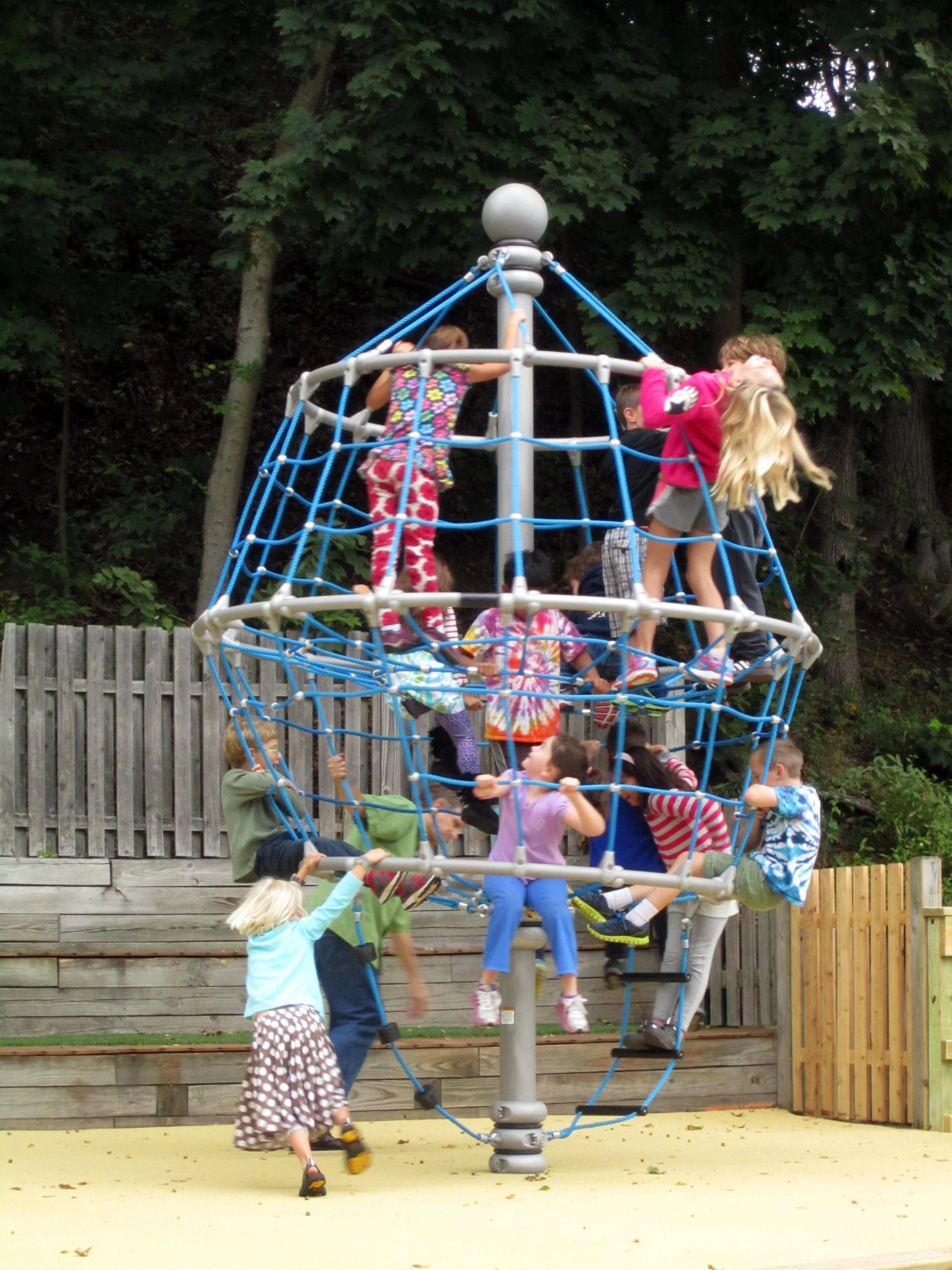 Children climb and play on a large rope climbing structure in an outdoor playground surrounded by wood fencing and mature trees.