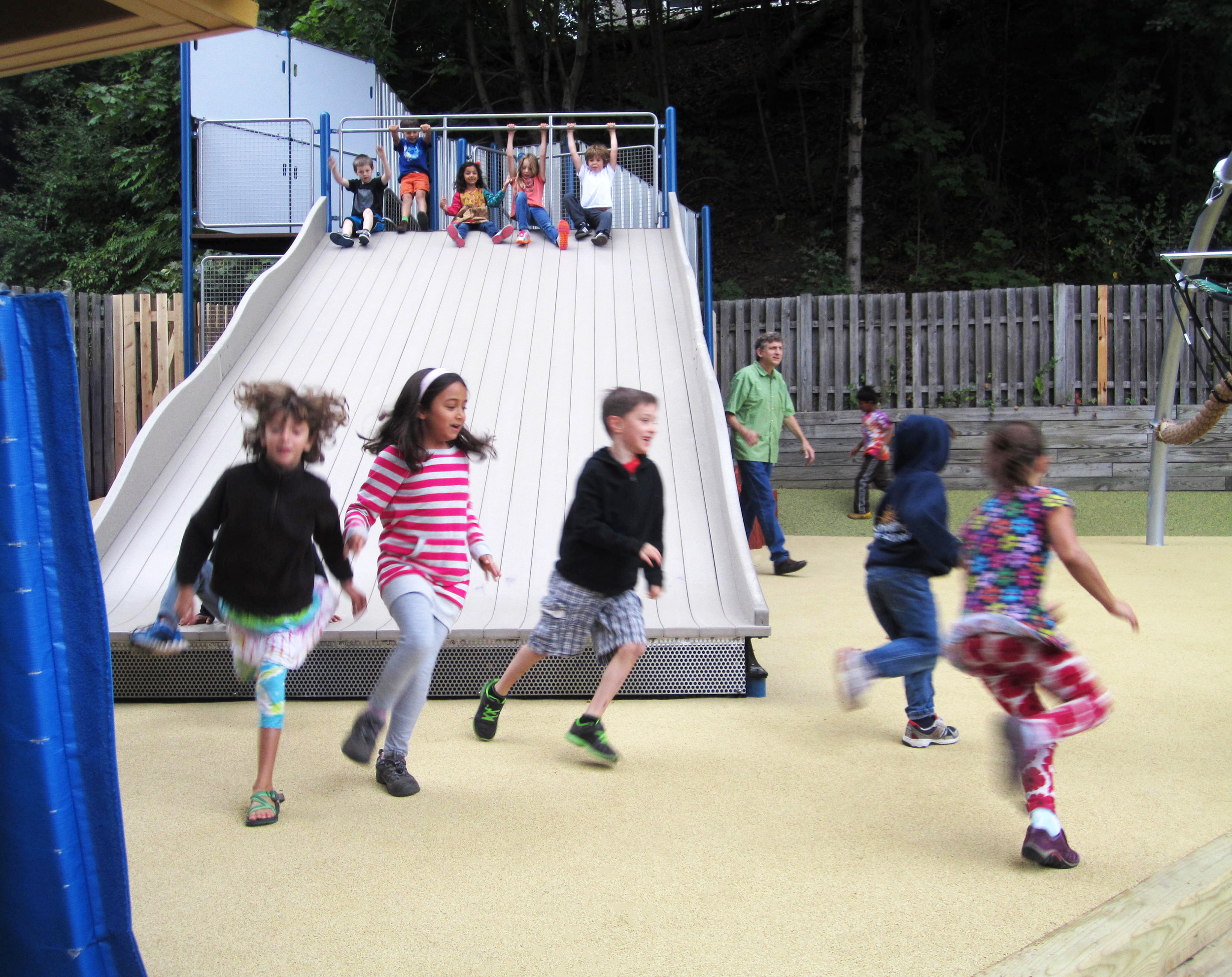 Children running and playing on a large custom playground slide with climbing equipment and wooded surroundings.