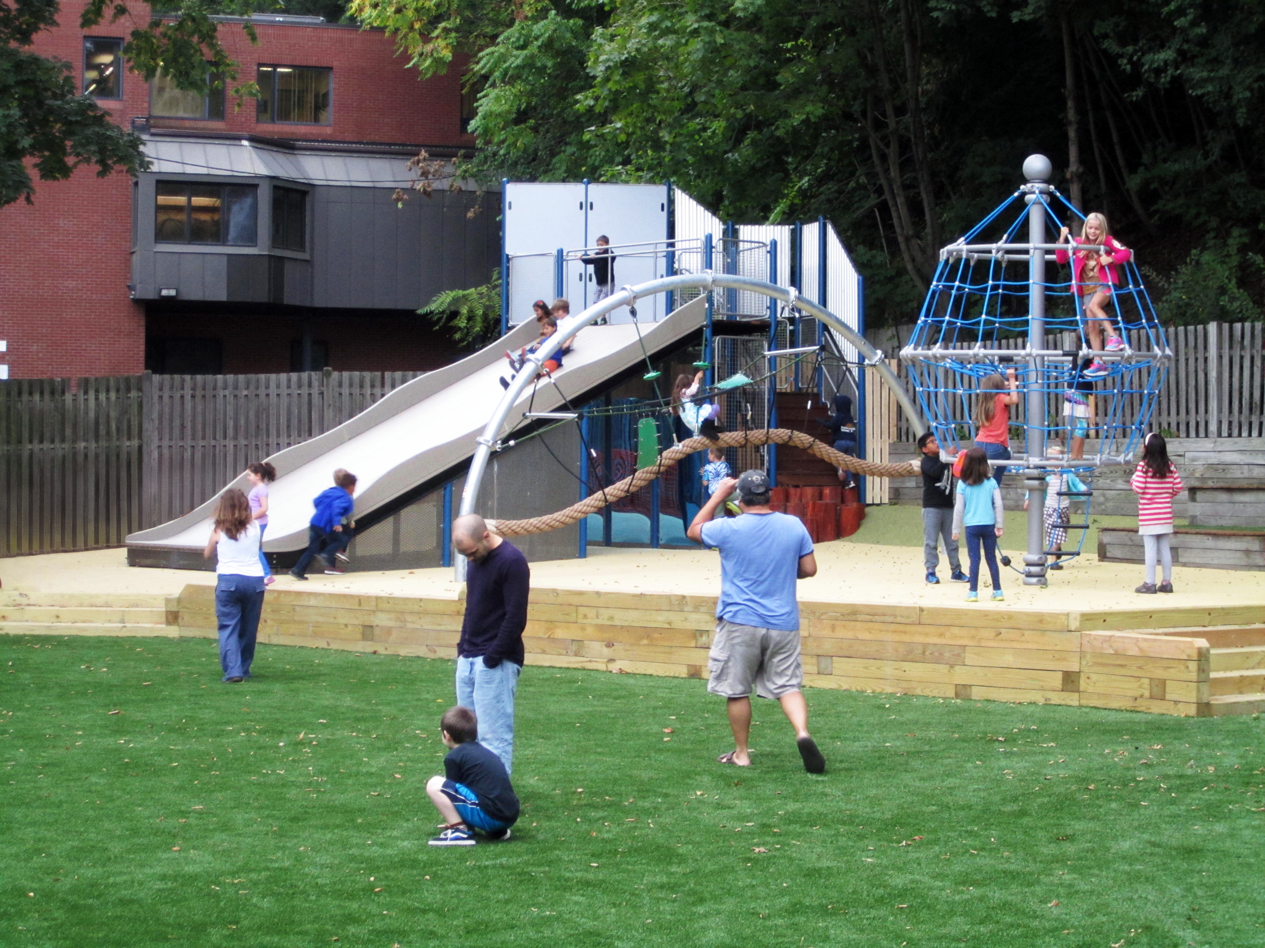 Children and adults using a school playground with a large slide, rope climbing structures, and an open grassy play area.