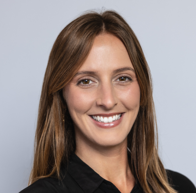 A person with long light brown hair wears a black collared shirt, smiling toward the camera against a light gray background.