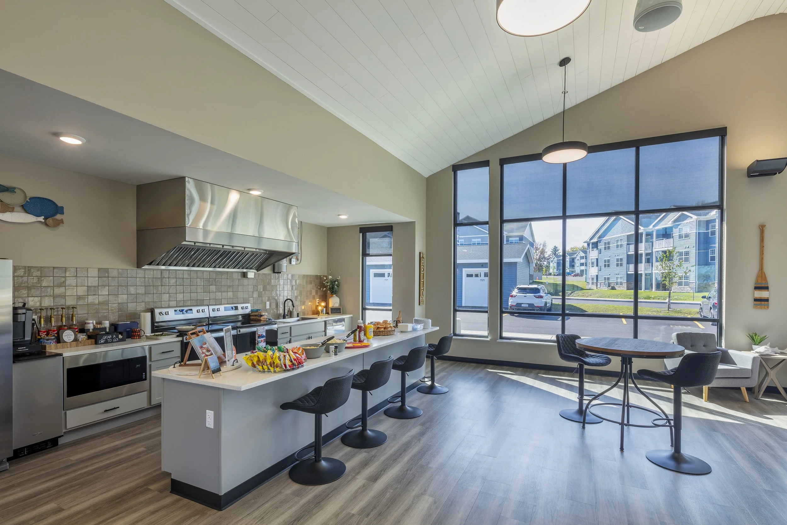 Community kitchen and gathering space featuring a large island with seating, café tables, high ceilings, and floor-to-ceiling windows overlooking a residential community.