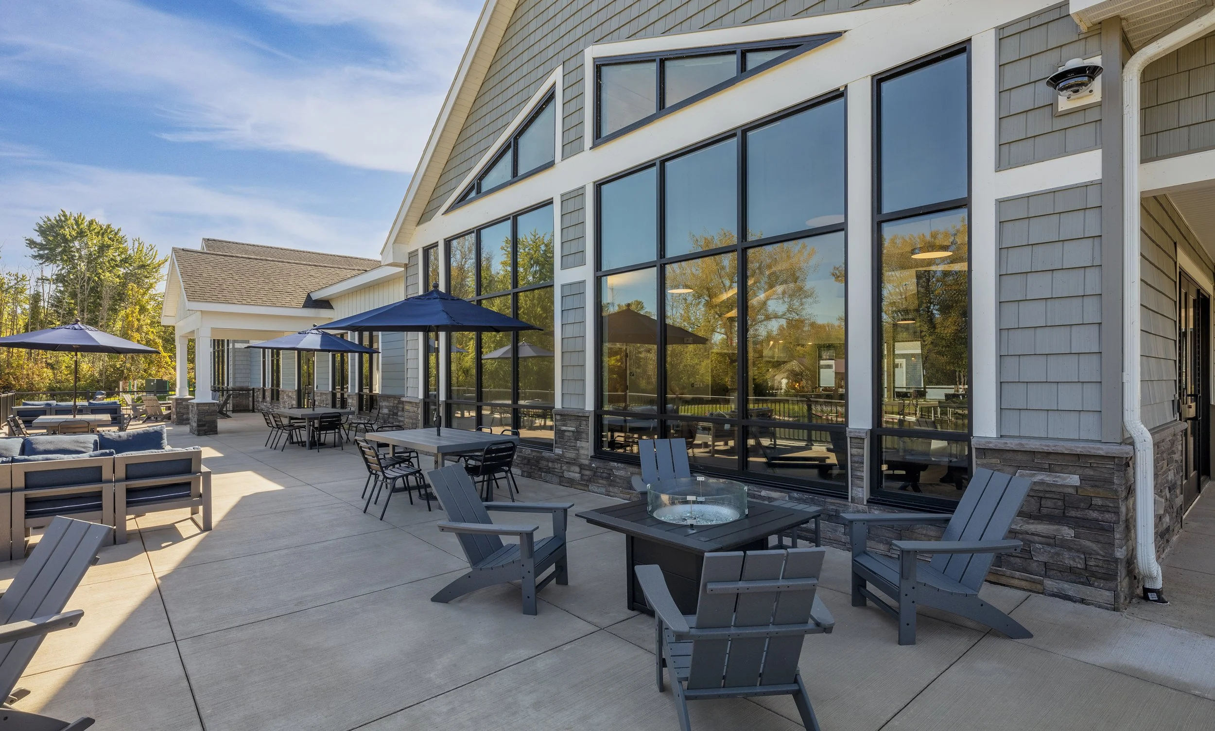 Outdoor patio featuring lounge seating, dining tables, umbrellas, Adirondack chairs, and a fire pit adjacent to a residential clubhouse.