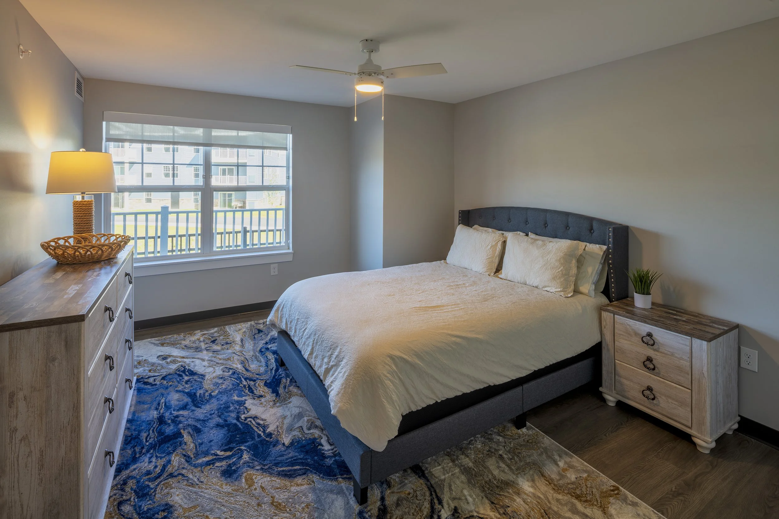 Bedroom featuring a queen-sized bed, nightstand, dresser, area rug, and large window overlooking a residential community.