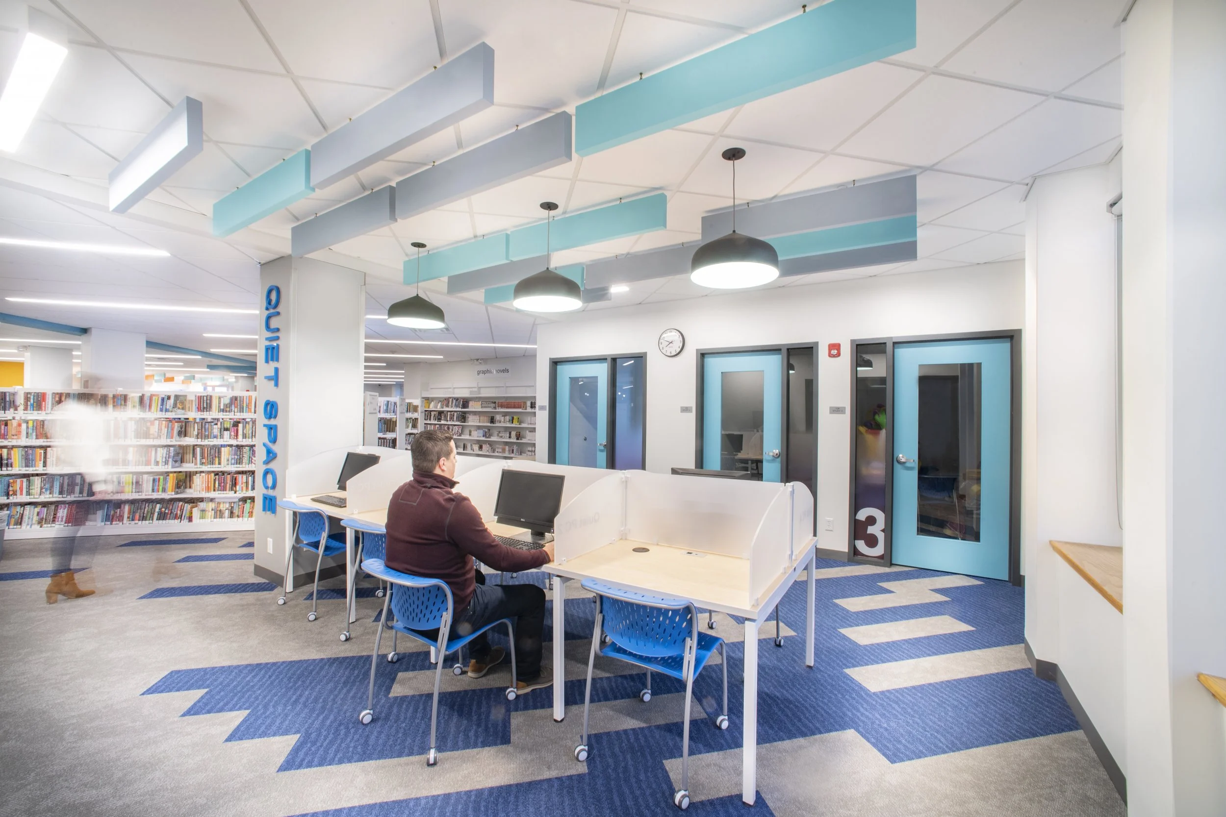 Open library study area with individual workstations, enclosed quiet rooms, bookshelves, and suspended acoustic panels.