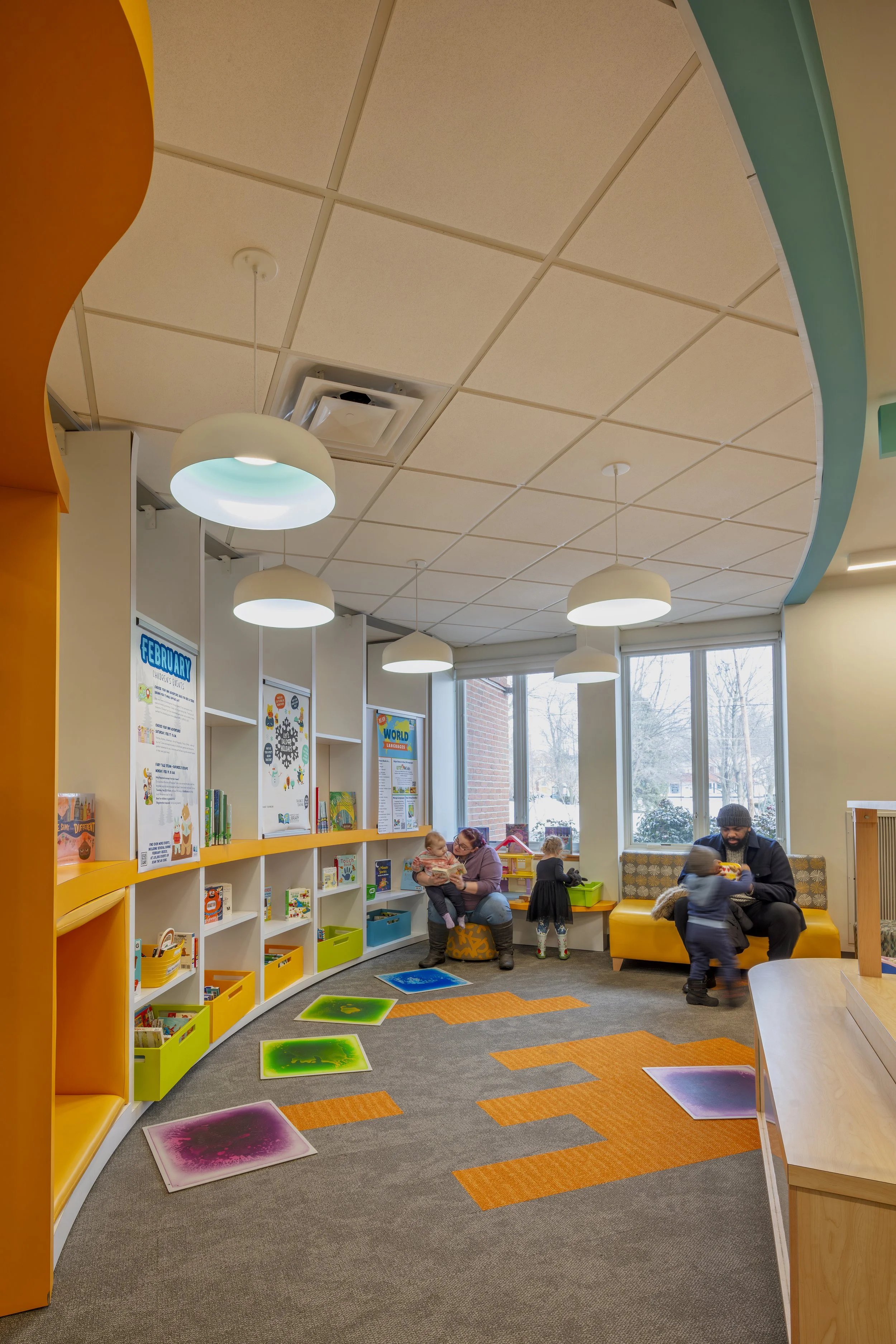 Families gather in a colorful children’s library area with books, seating, and sensory mats on the floor.