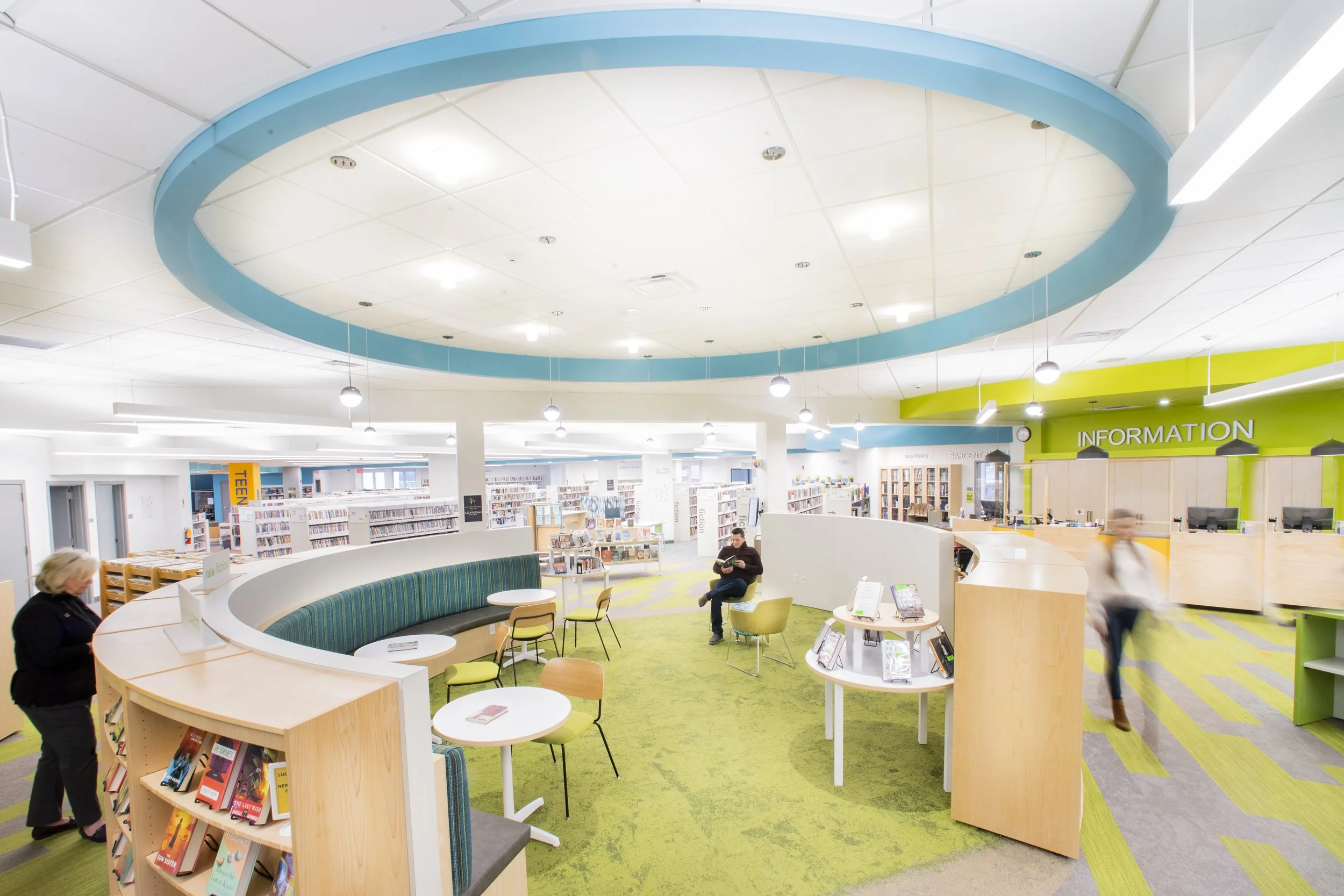 Open library interior with curved seating, an information desk, bookshelves, and a large circular ceiling feature.