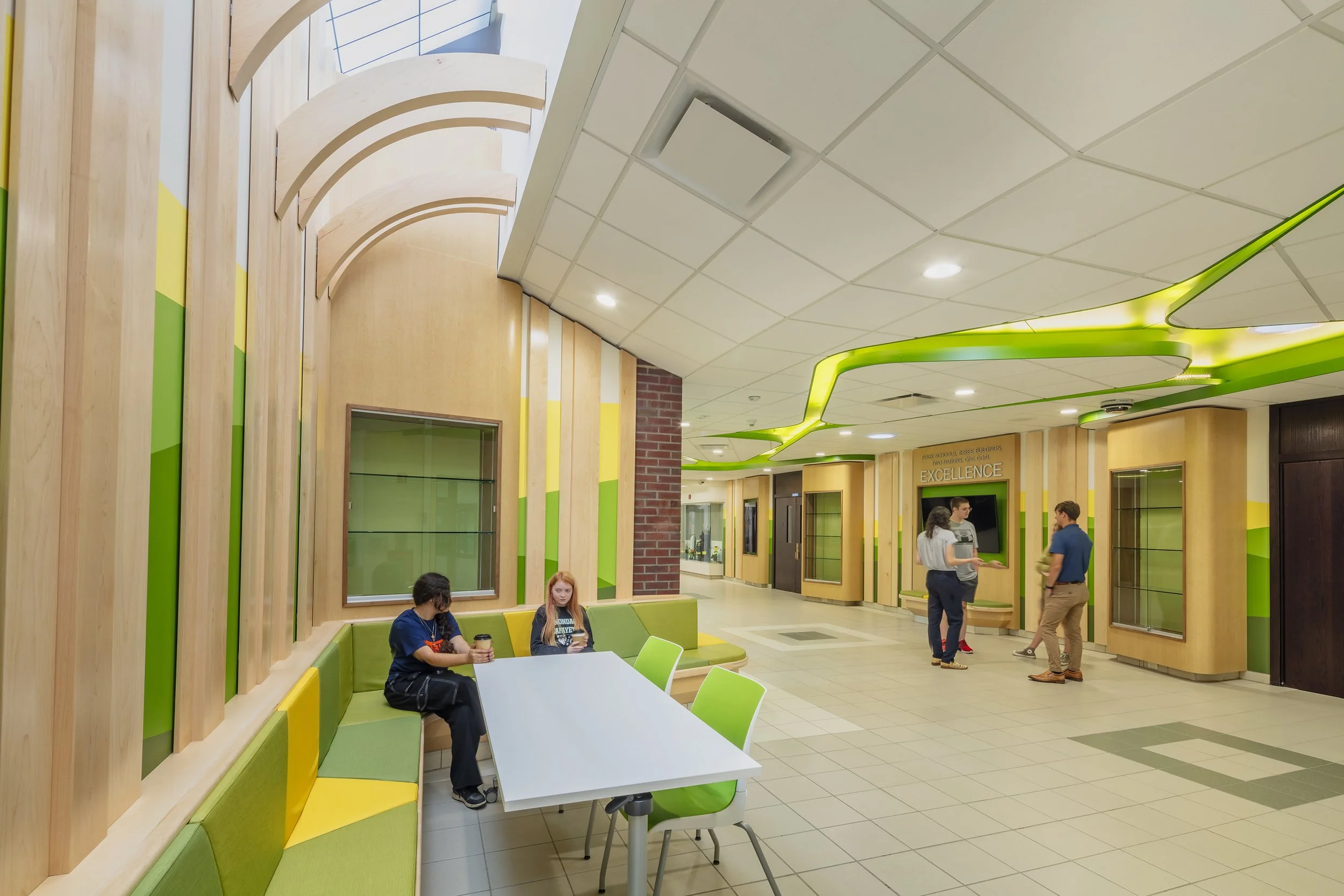Students seated in a collaborative learning area featuring green and yellow upholstered benches, flexible tables, and natural wood architectural details.