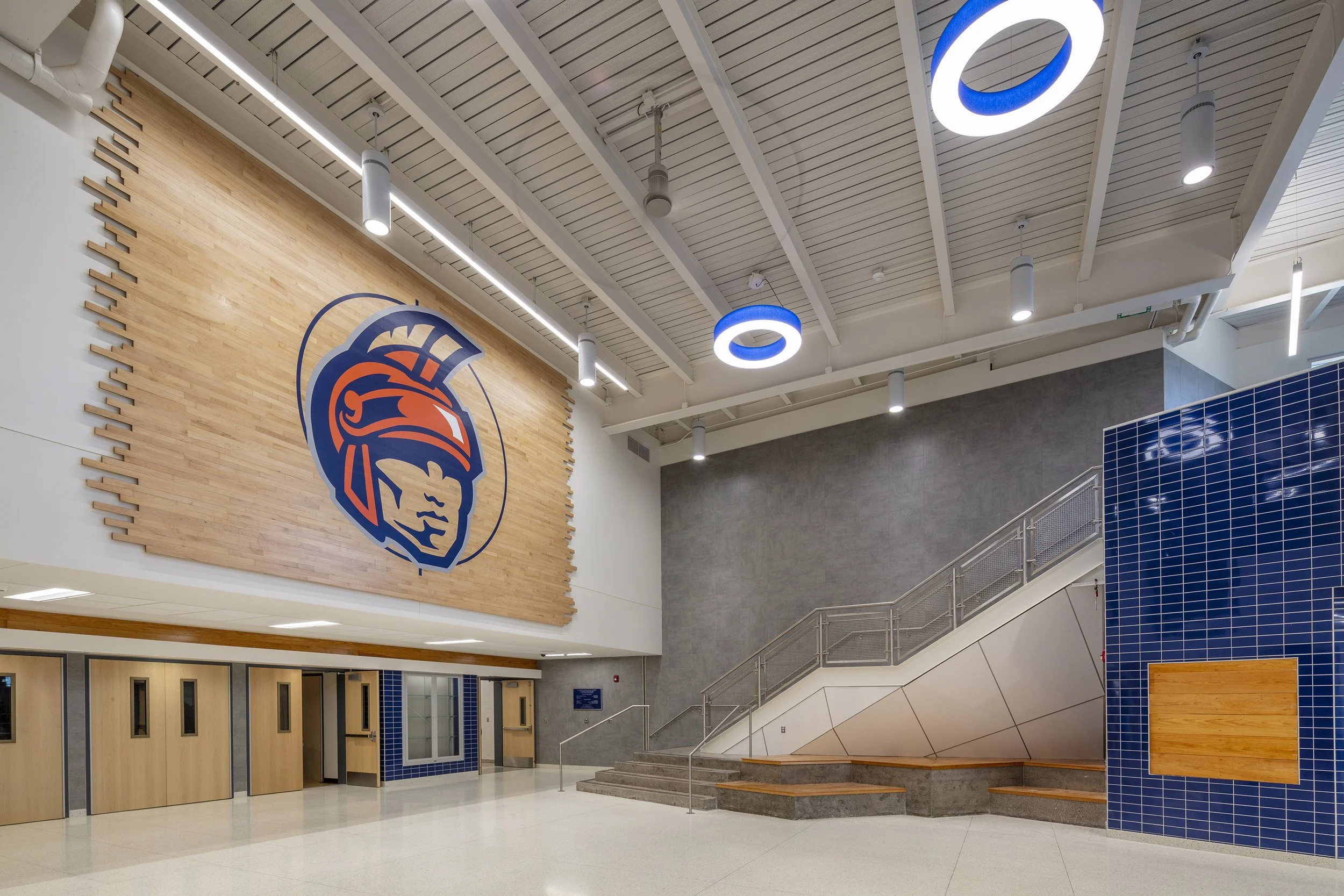 Liverpool High School athletics lobby with Warriors branding, wood accent wall, blue tile, integrated seating, and central staircase.