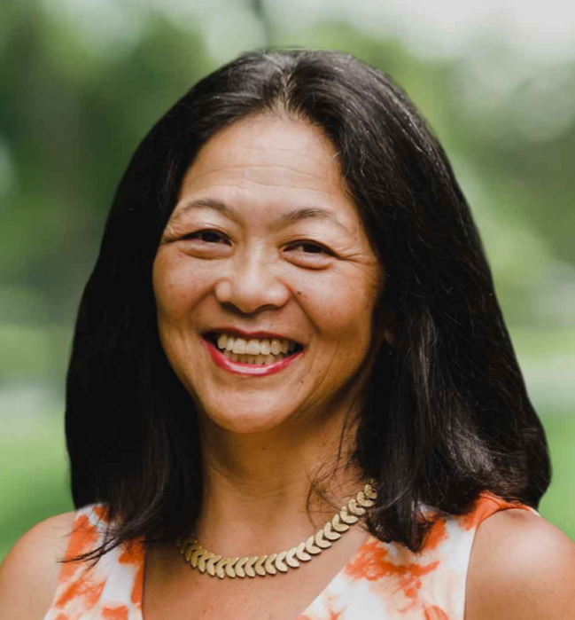 A person with shoulder-length black hair wears an orange and white patterned sleeveless top and a gold necklace, smiling toward the camera against a softly blurred green outdoor background.