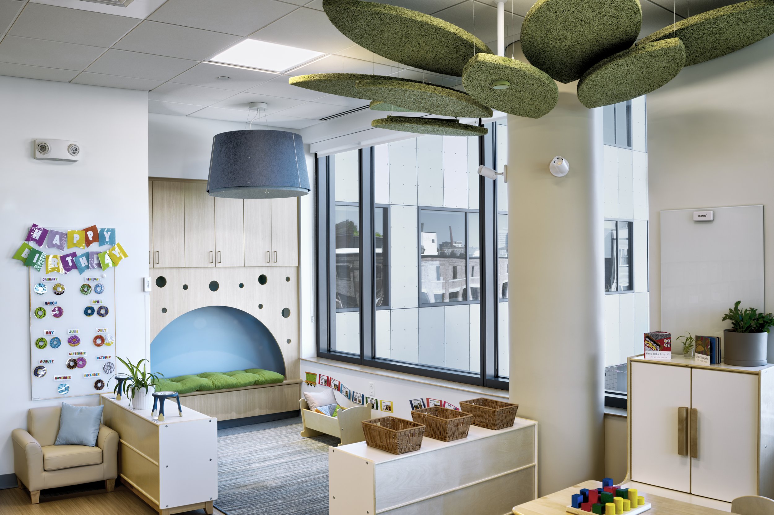 Bright classroom with floor-to-ceiling windows, a semi-enclosed reading nook, child-scaled furnishings, and biophilic design elements that promote comfort and connection to nature.