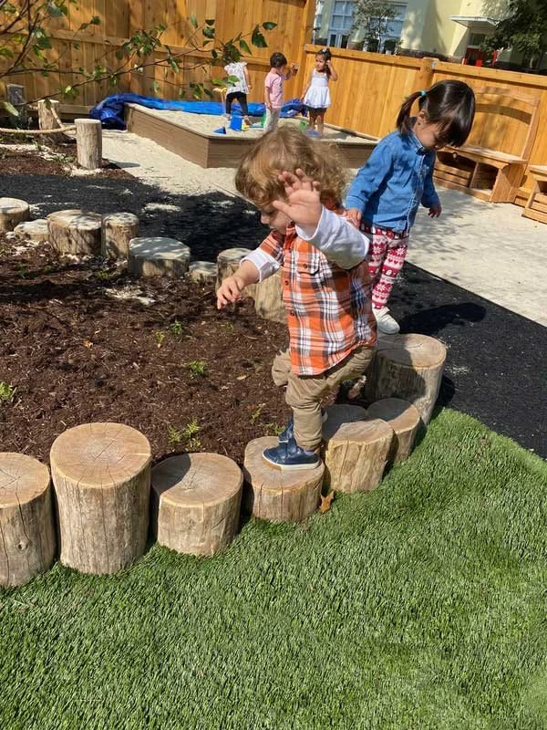 Young children balance across natural wood stepping stumps in an outdoor play area designed to encourage movement, coordination, and exploration.