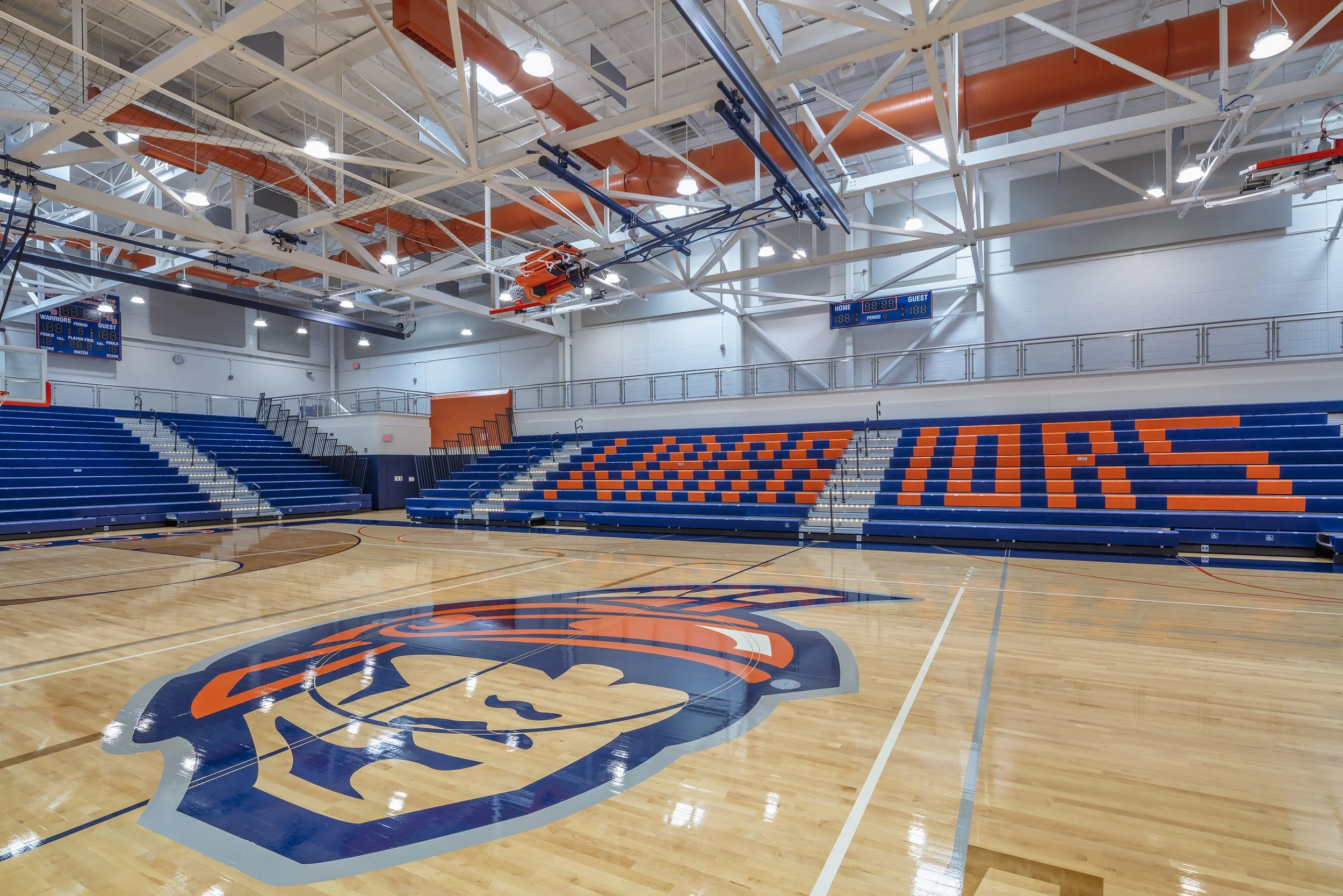 Liverpool High School gymnasium featuring the Warriors logo at center court and blue and orange bleacher seating.
