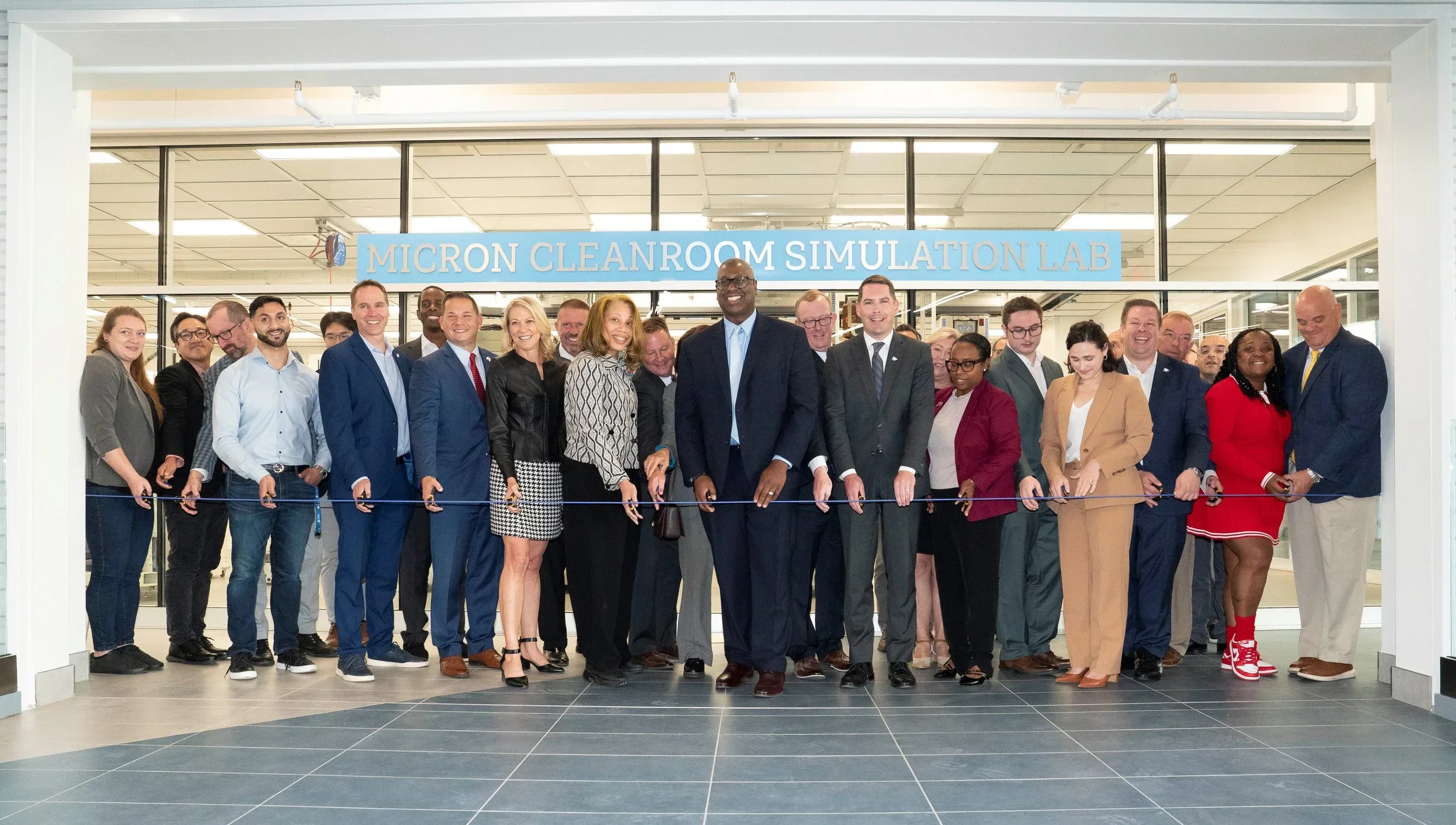 A large group of community leaders, educators, and industry partners participate in a wire-cutting ceremony celebrating the opening of the Micron Cleanroom Simulation Lab, standing in front of the facility entrance with ceremonial scissors.