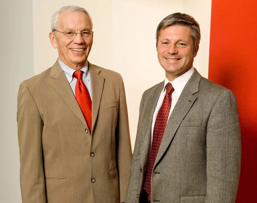 Two men in suits standing and smiling in front of a wall with red, white, and beige colors.